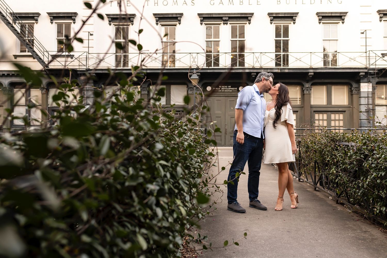 A couple shares an intimate moment together along a path lined with ornate architecture and greenery in Paris.