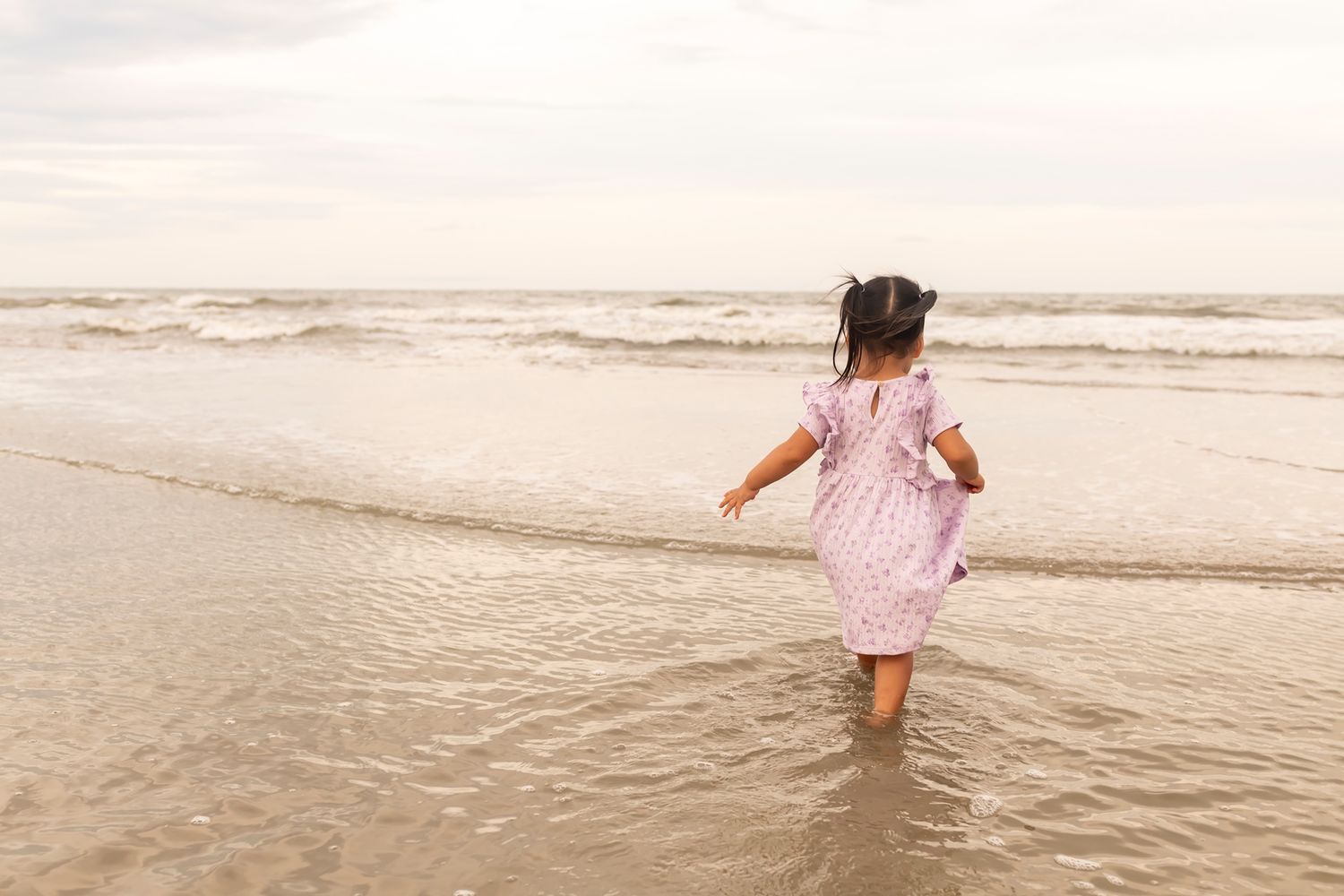 A small child in a pink dress wades through shallow ocean waves on a sandy beach at sunset.