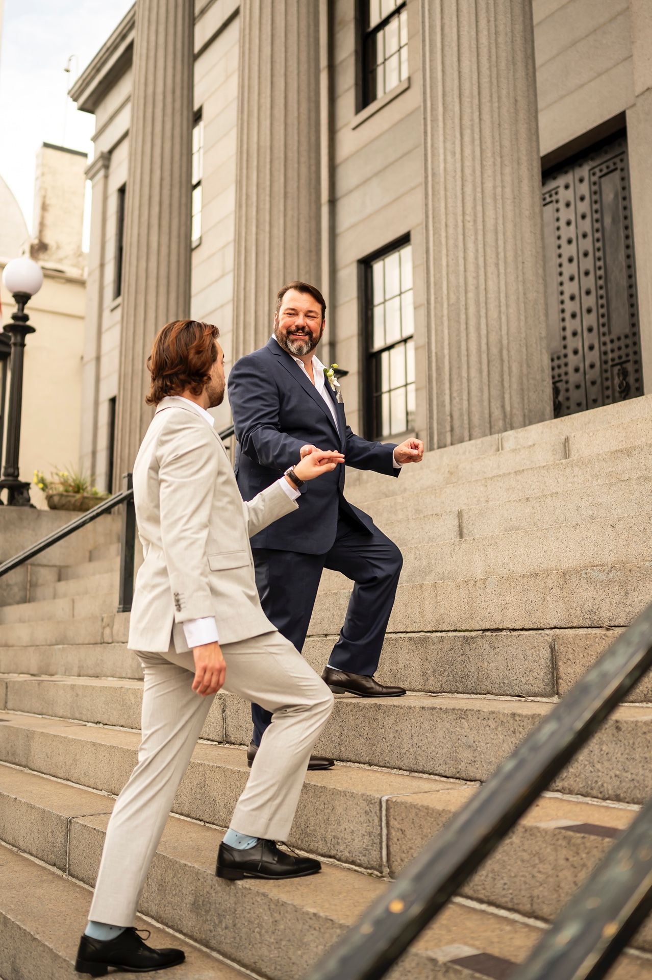 Two people in formal attire playfully interact on stone steps outside a neoclassical building with columns.
