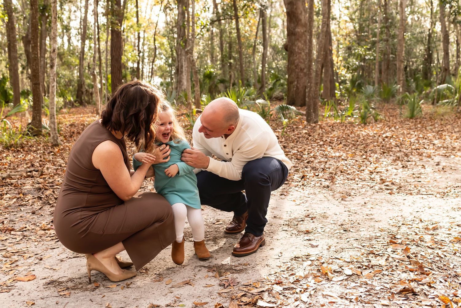 Three people crouch on a leaf-covered forest path while playing together in warm afternoon sunlight.