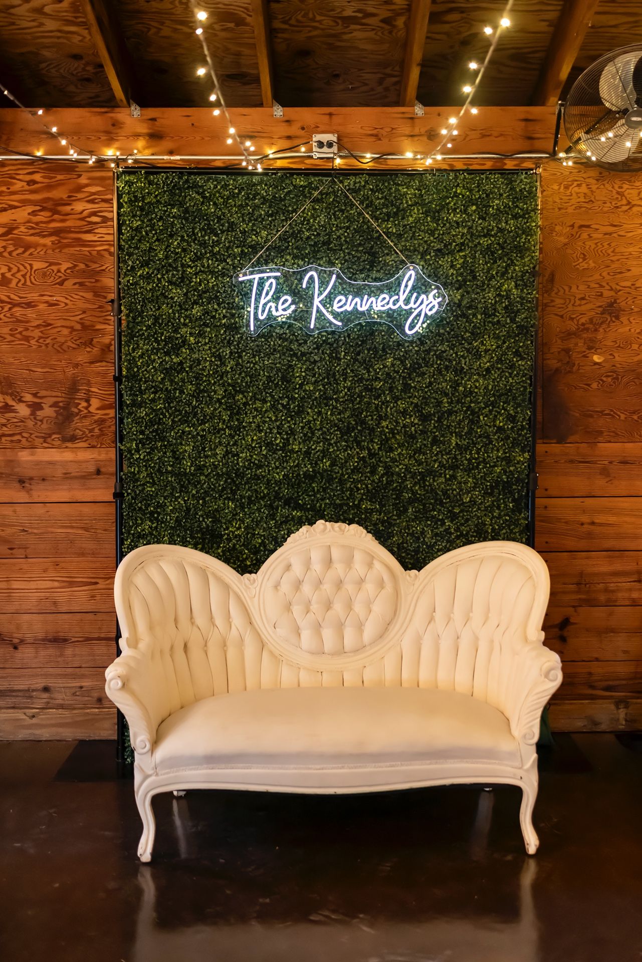 Vintage white Victorian sofa against green hedge wall with neon sign reading 'The Knowledge' in a rustic wooden venue.