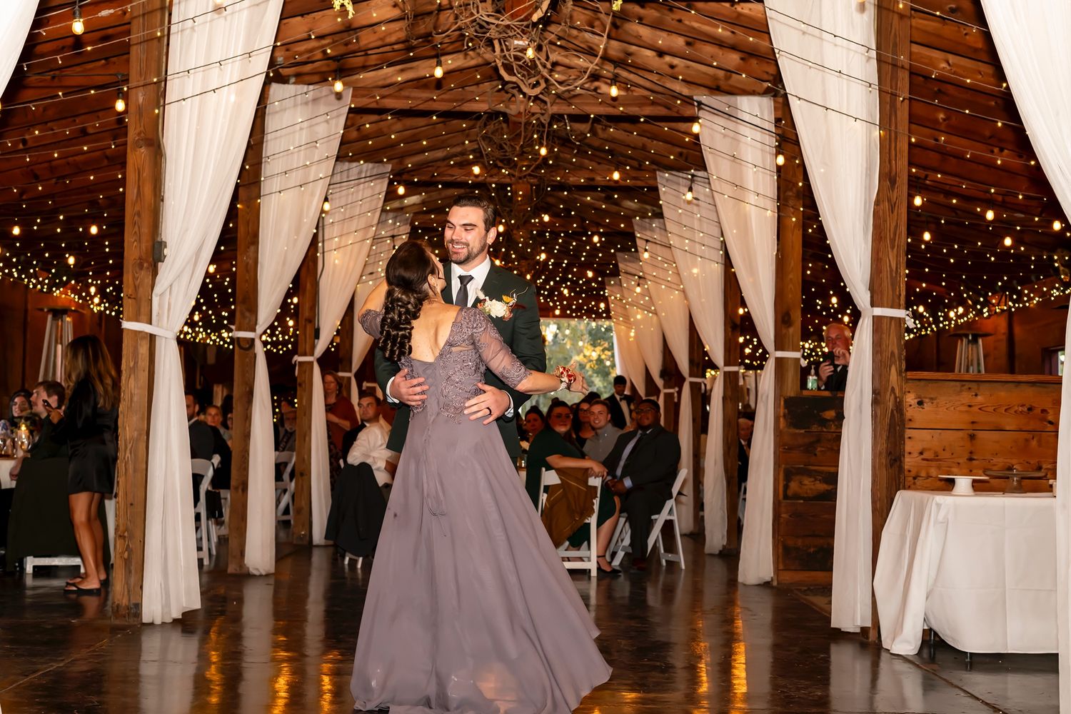 A couple dances together during a wedding reception in a rustic barn venue illuminated with string lights.