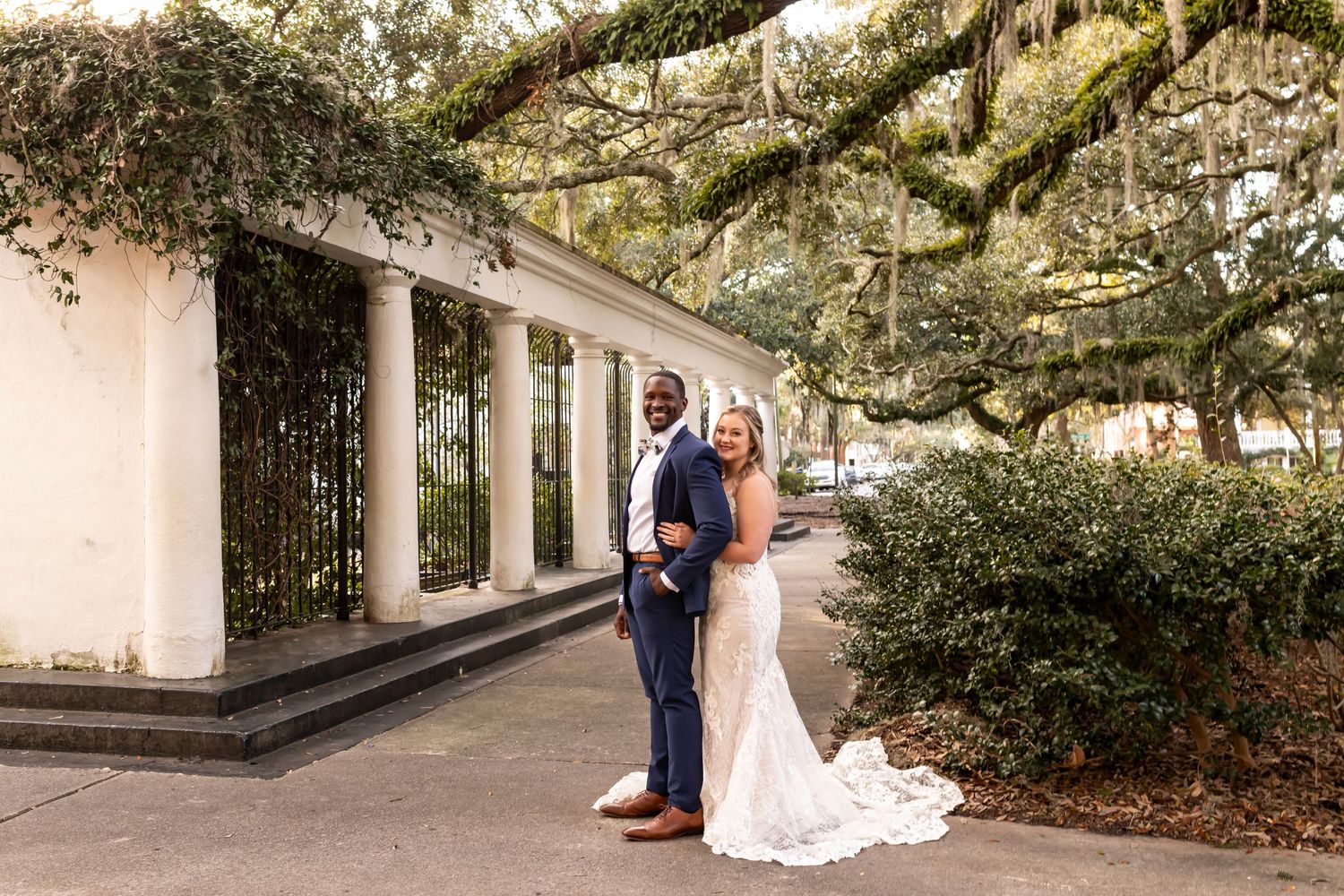 A happy couple stands beneath Spanish moss-draped trees by white columns at a scenic outdoor venue.