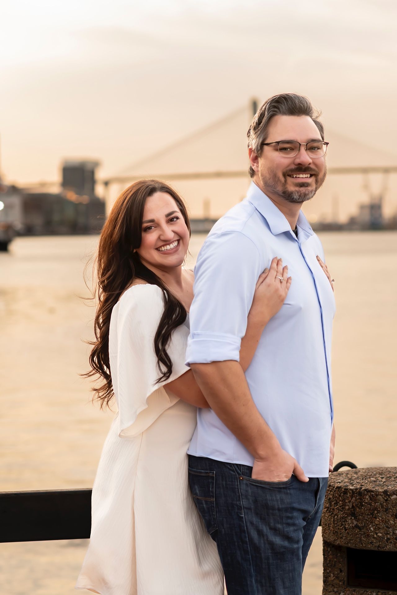Couple poses together at sunset near a waterfront with a bridge visible in the background.