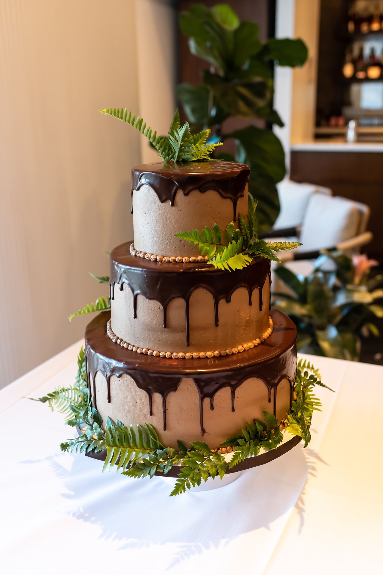 Three-tiered white wedding cake decorated with chocolate ganache drips and green fern leaves on a white tablecloth.