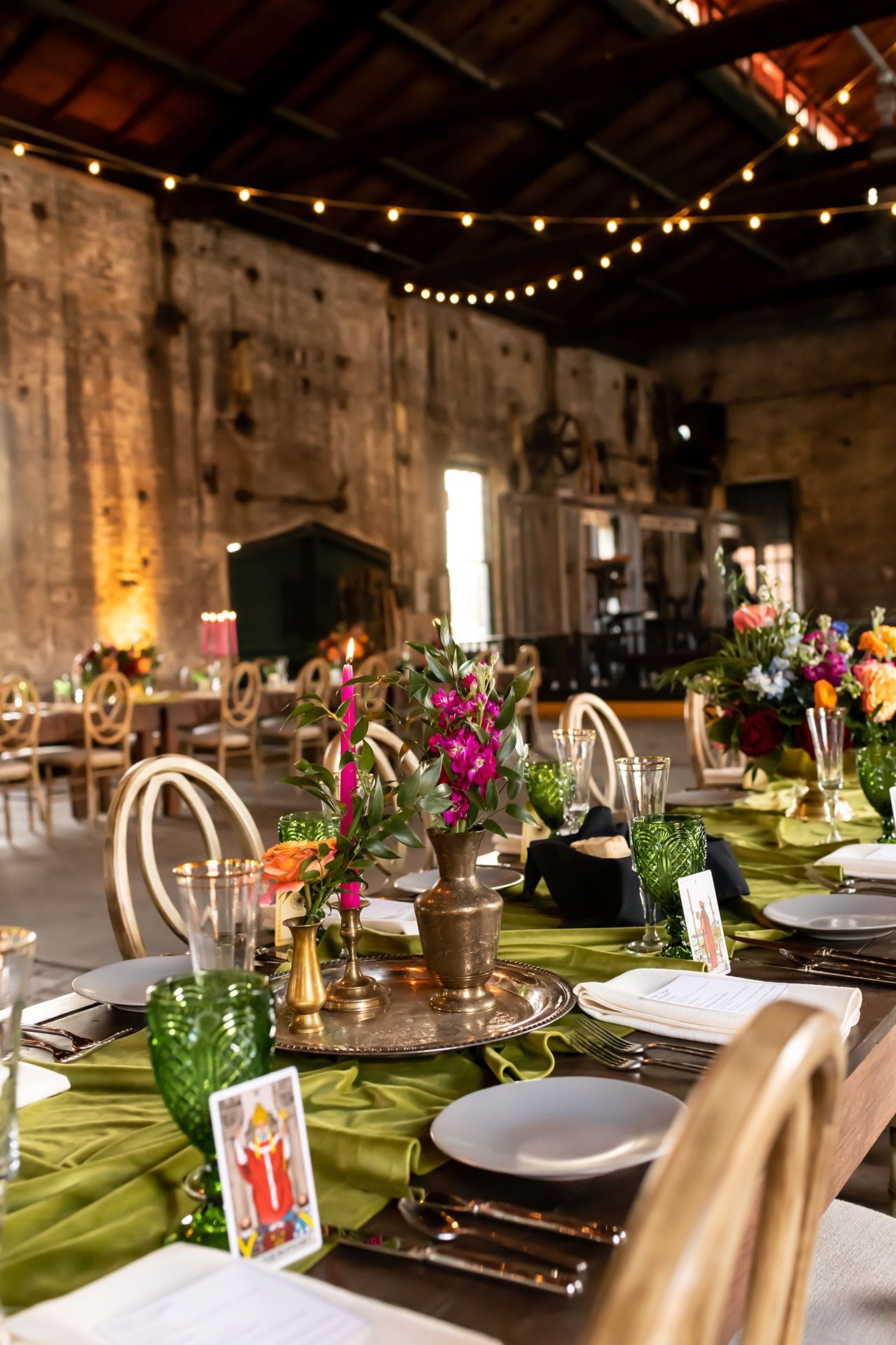 Rustic barn wedding reception table with string lights, green tablecloths, and colorful floral centerpieces in mason jars.