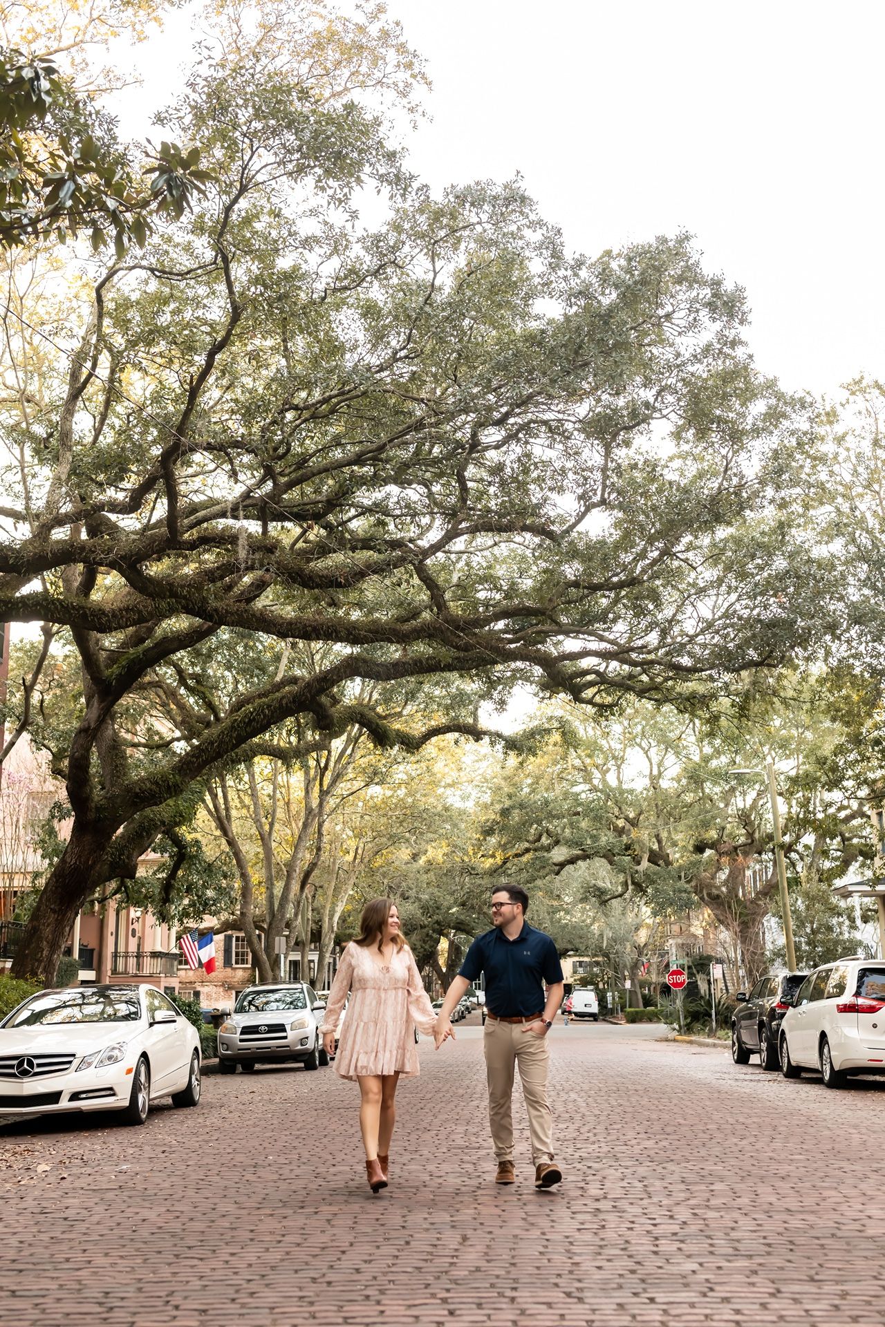 A couple strolls down a charming brick-paved street lined with Spanish moss-draped oak trees and parked cars in a historic district.