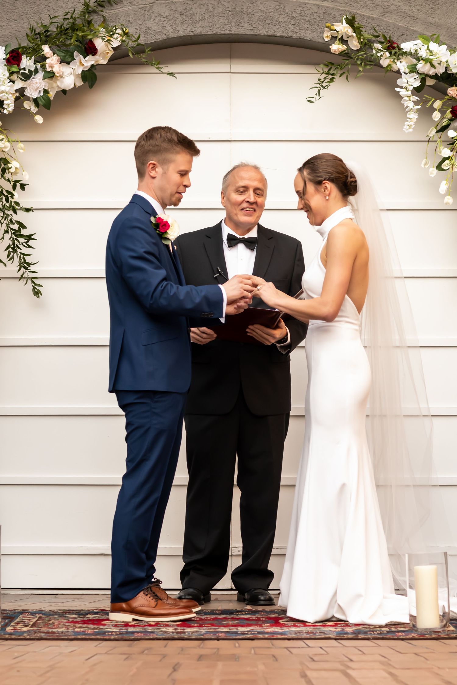 A couple participates in a wedding ceremony outdoors with an officiant on a patterned carpet.