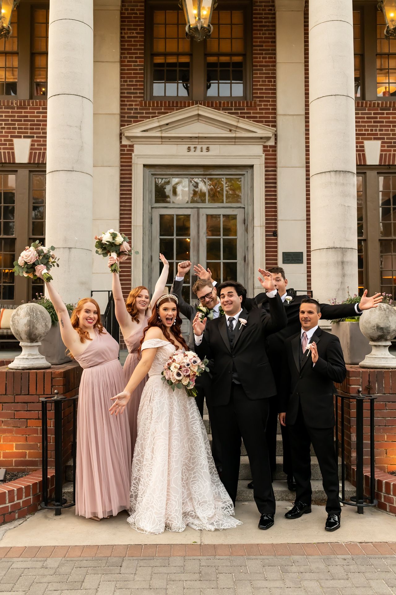 Wedding party poses with joy outside elegant brick building with white columns in coordinated blush and black attire.