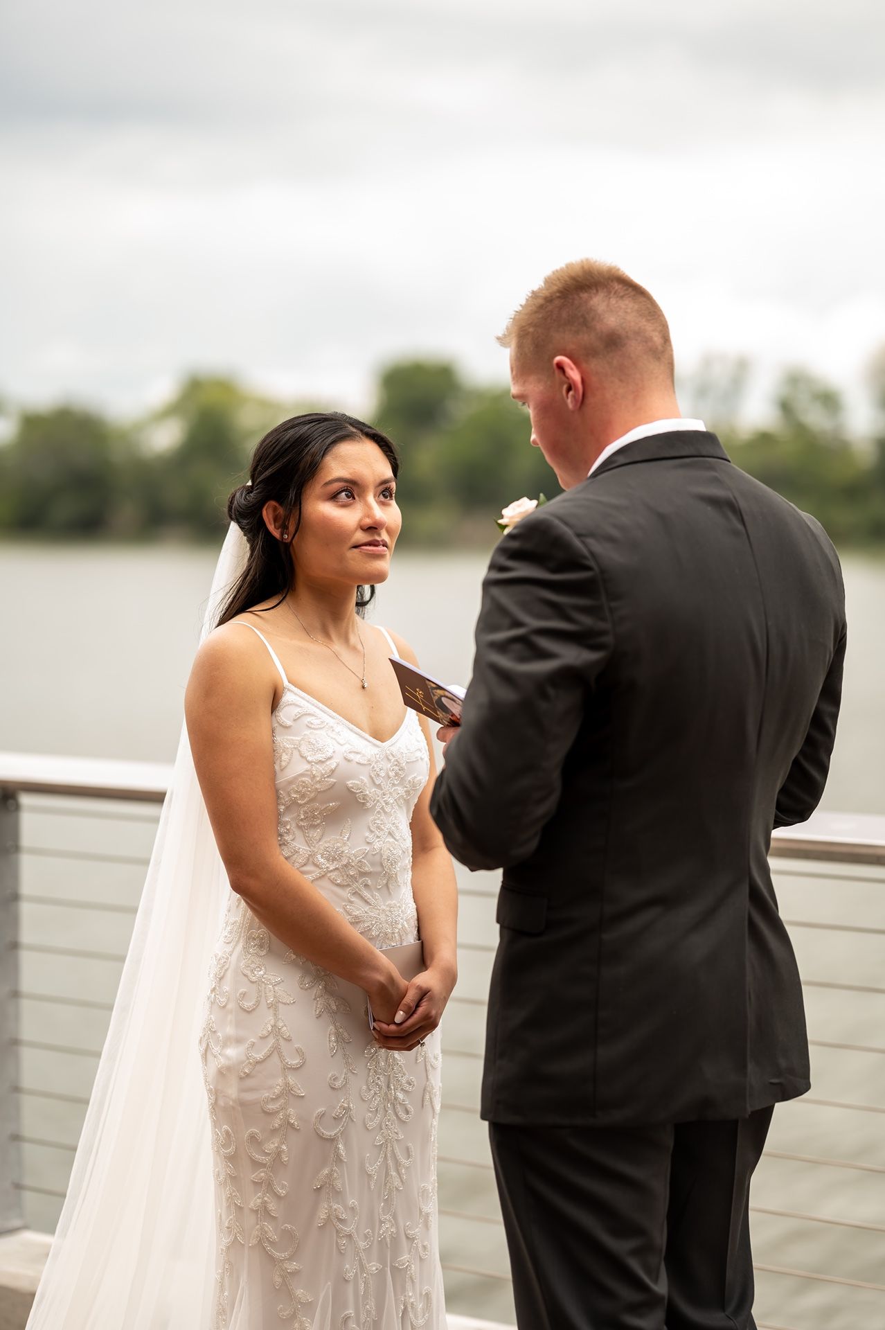 A bride in a lace wedding dress listens intently as her groom reads vows during their waterfront ceremony.
