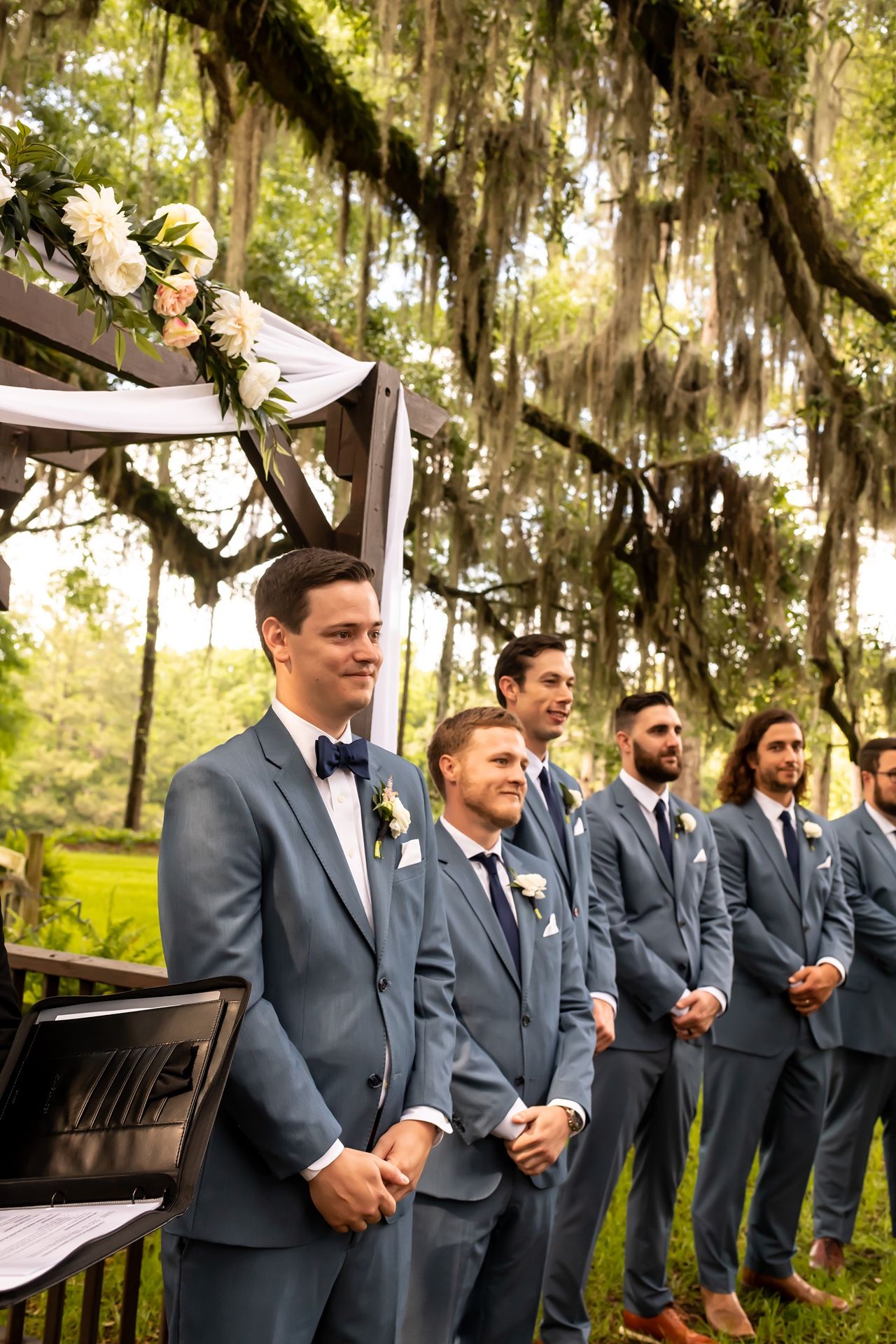 Groomsmen in matching gray suits stand beneath Spanish moss-draped oak trees at an outdoor wedding ceremony.