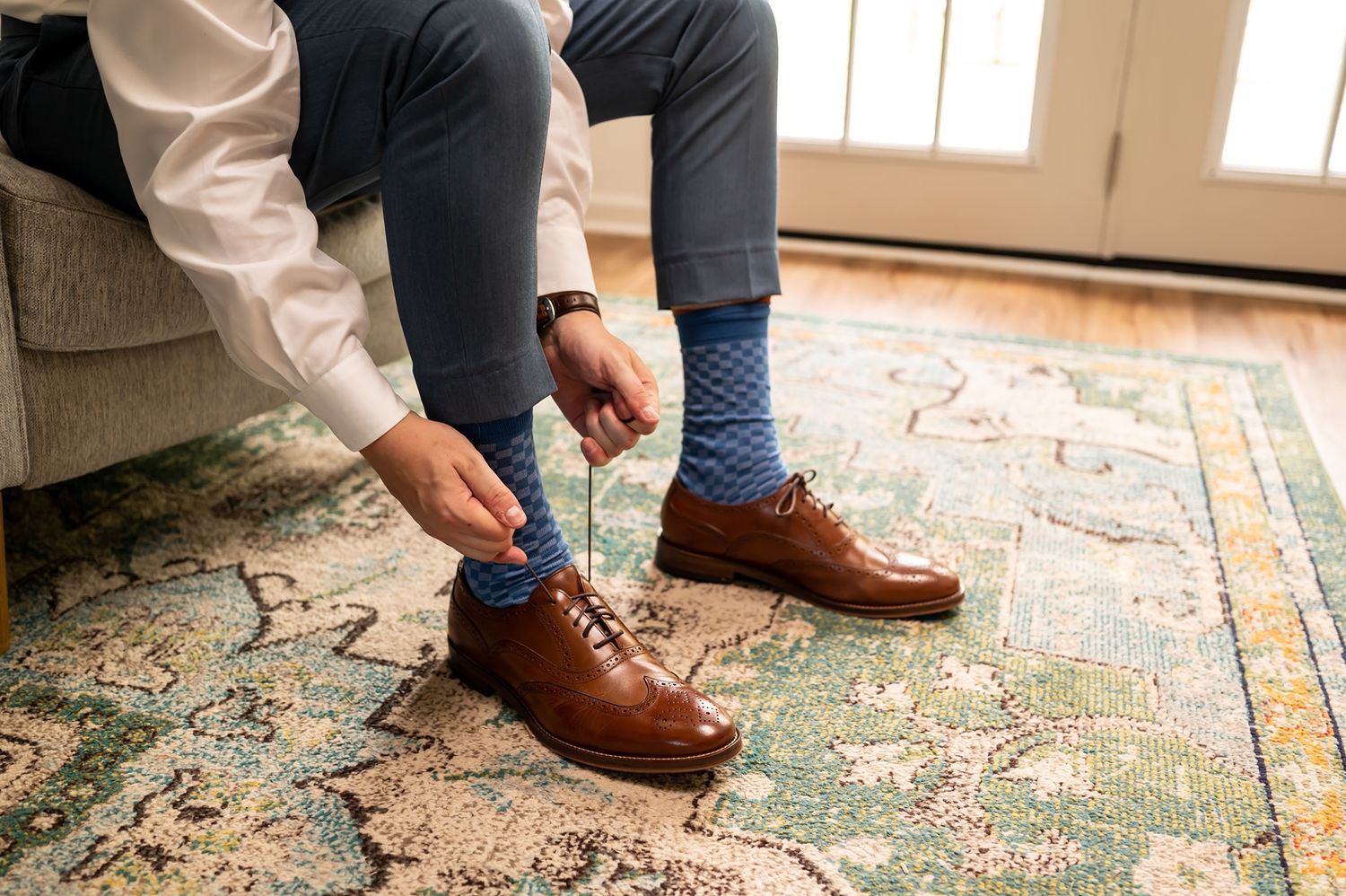 Person tying brown leather dress shoes while sitting on chair over decorative vintage rug.