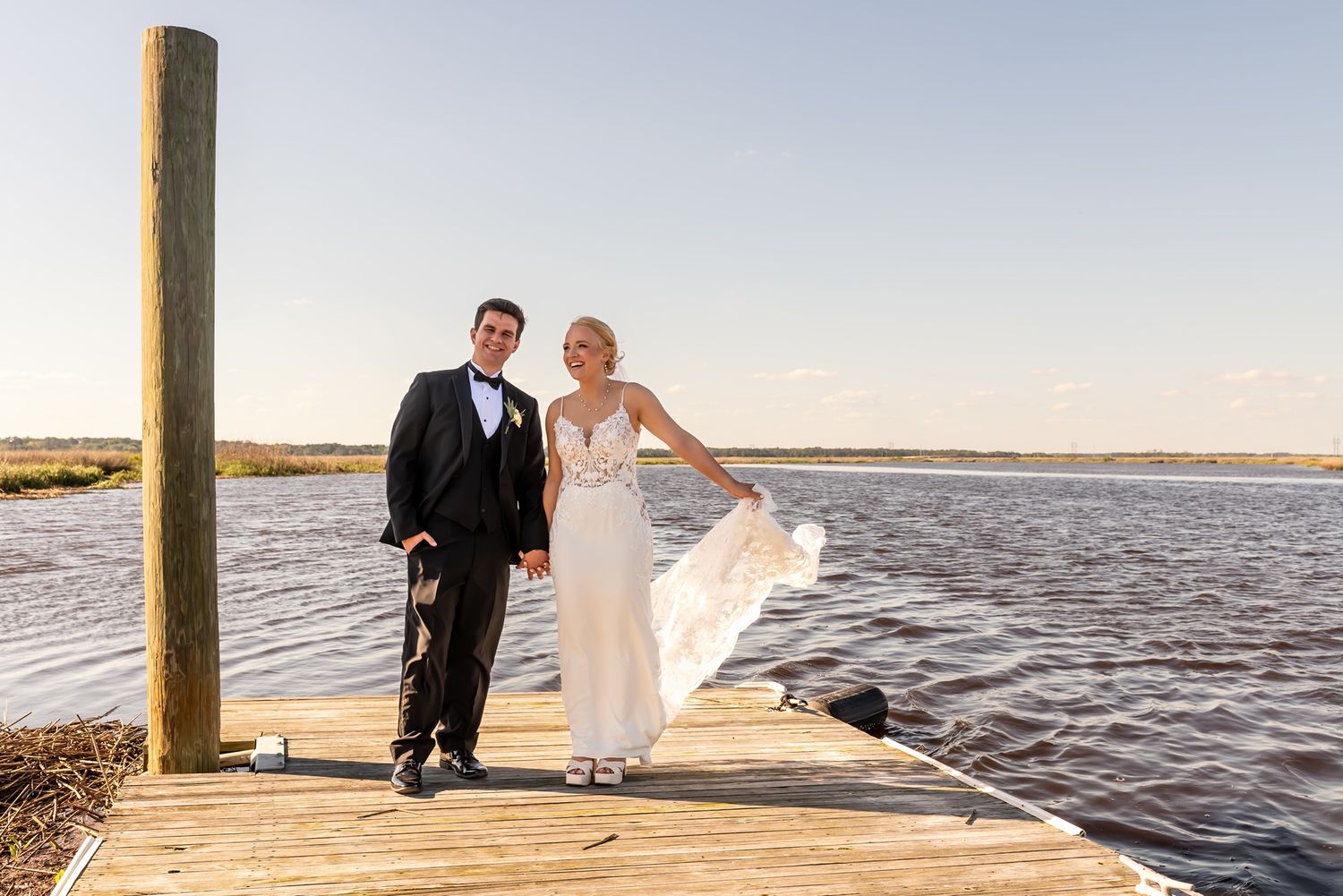 A couple poses on a wooden dock overlooking a scenic waterfront during sunset on their wedding day.