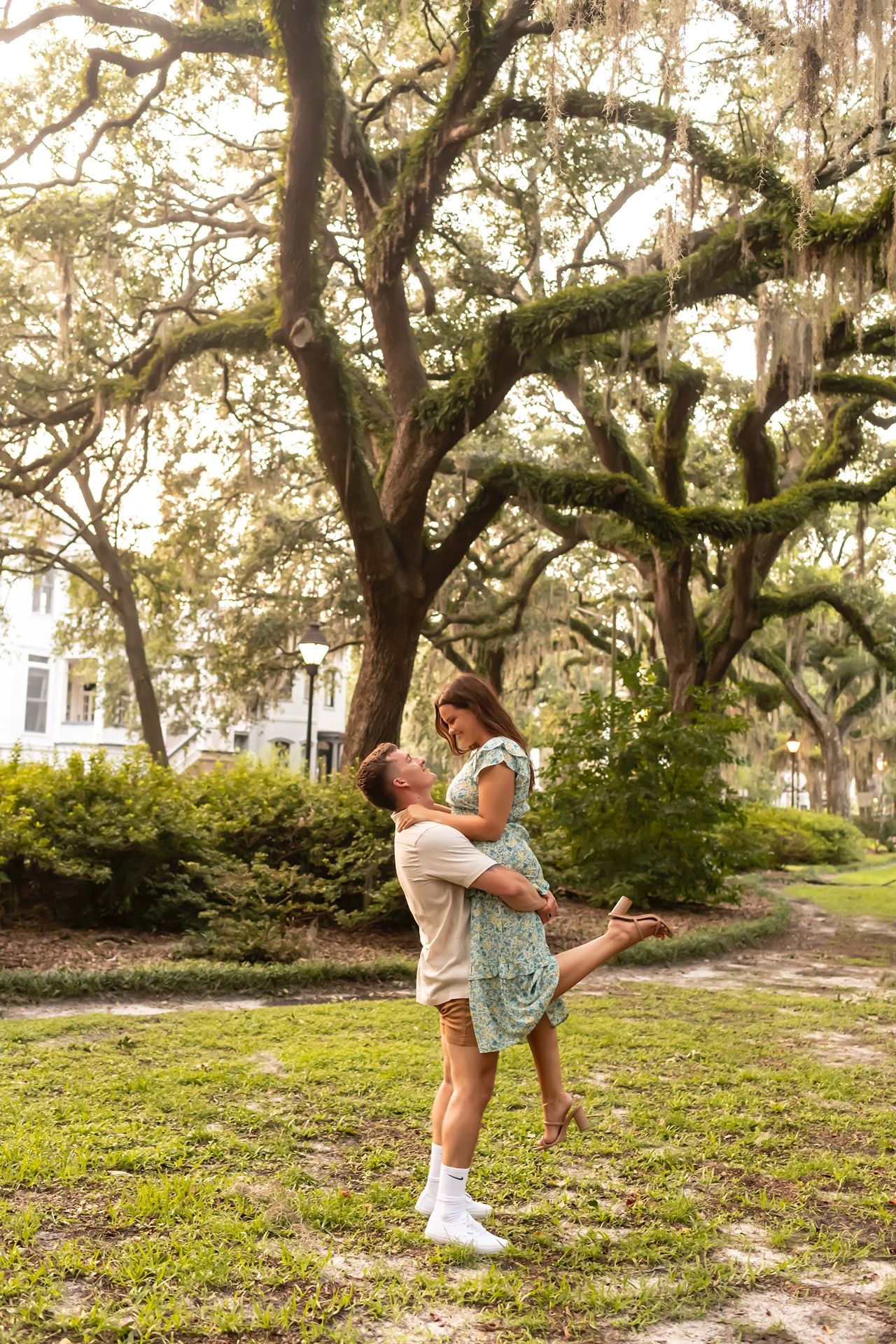 A couple embraces under Spanish moss-draped oak trees in a romantic park setting during golden hour.