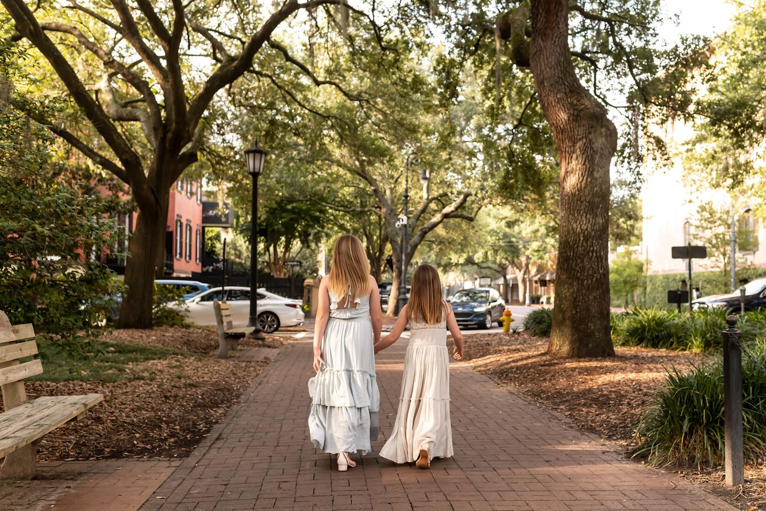 Two people in flowing dresses walk hand in hand down a tree-lined brick pathway in a scenic southern town.