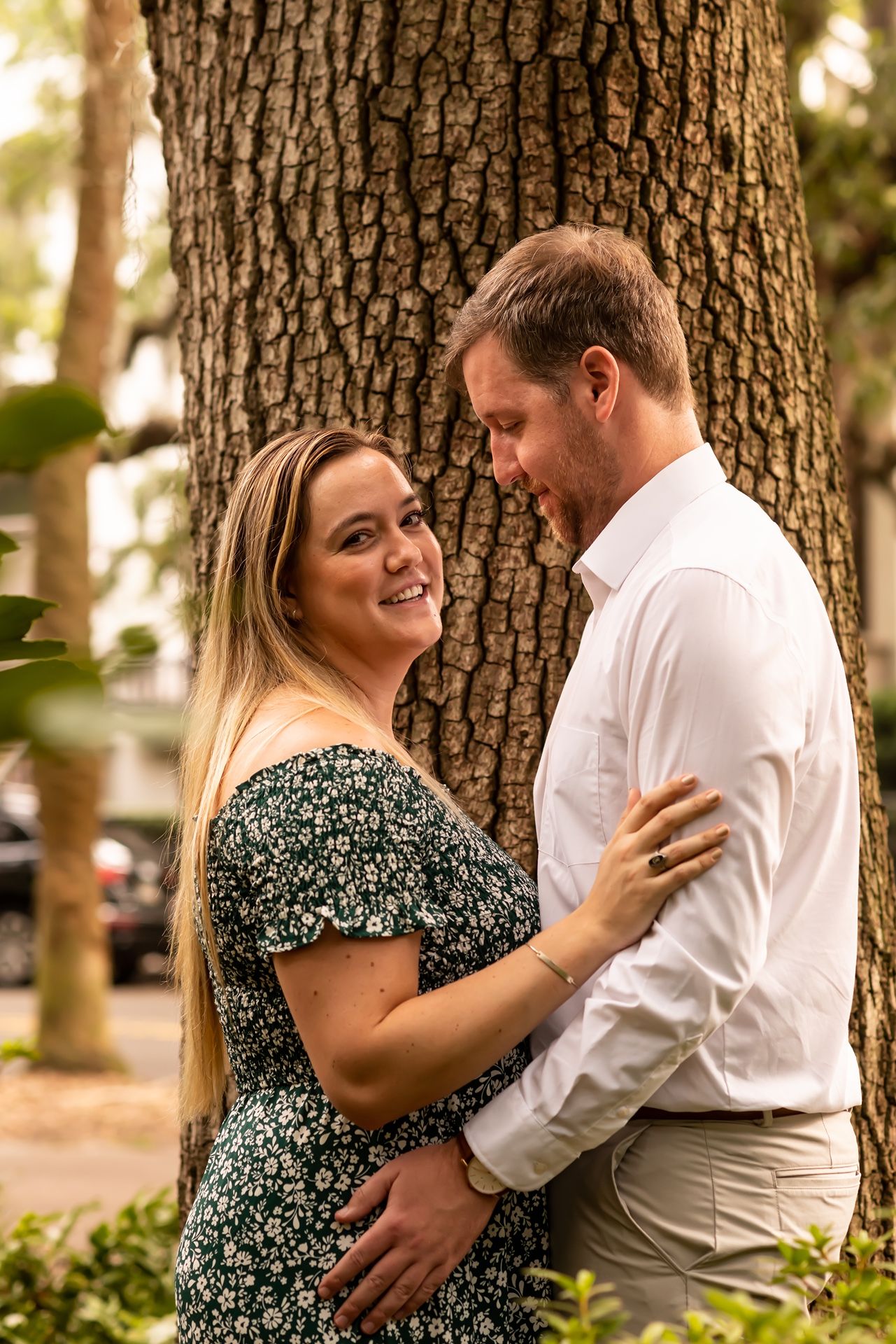 A couple shares an intimate moment against a large tree trunk in a romantic outdoor setting at golden hour.