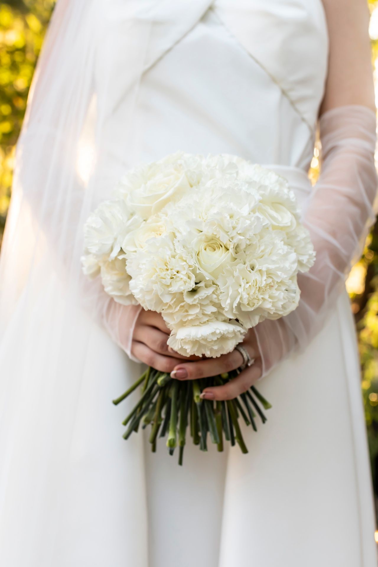 Close-up of bride holding classic white peony and carnation wedding bouquet against ivory dress.