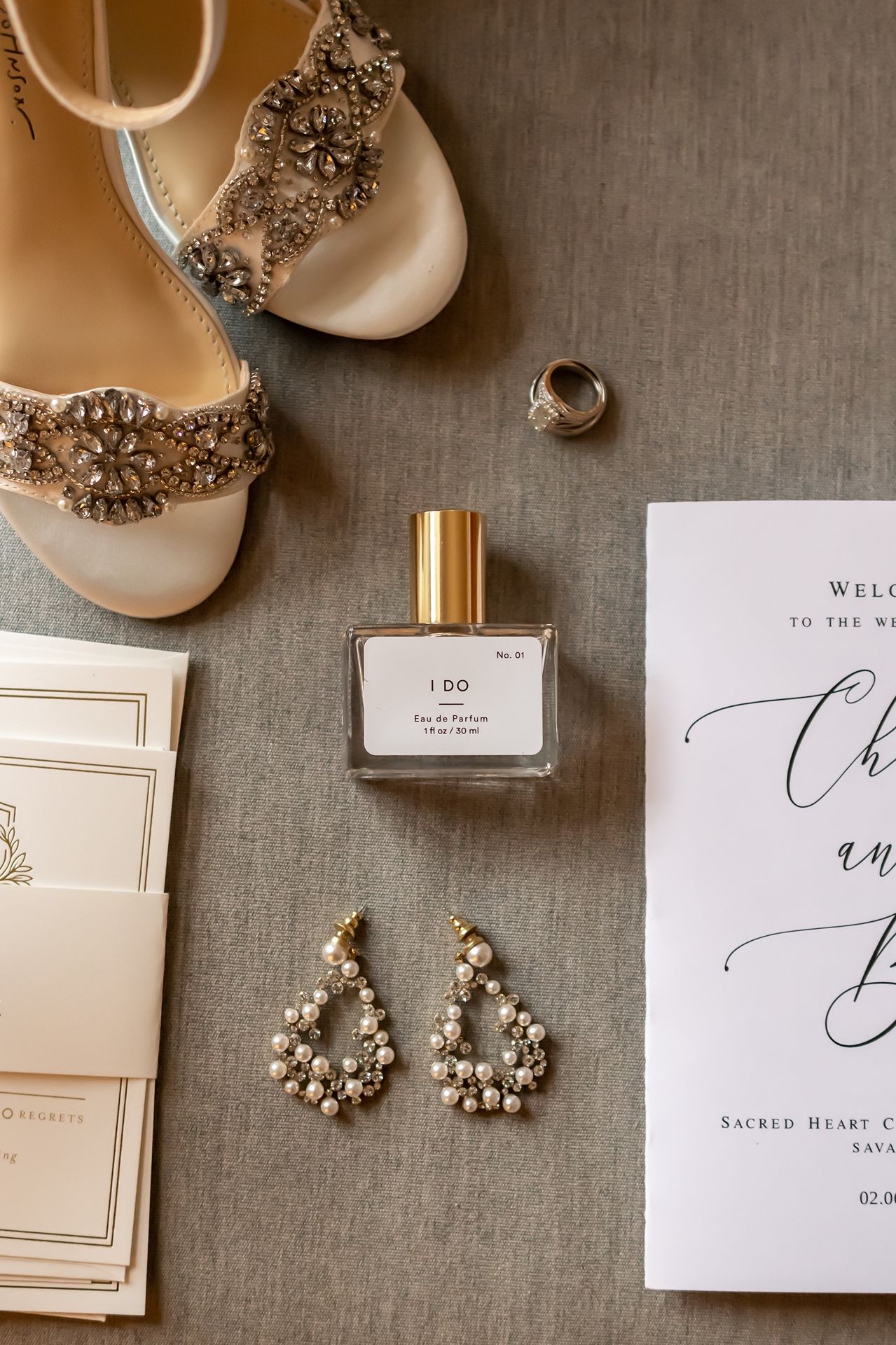 Wedding accessories including embellished shoes, perfume, invitation and crystal earrings arranged on grey linen.