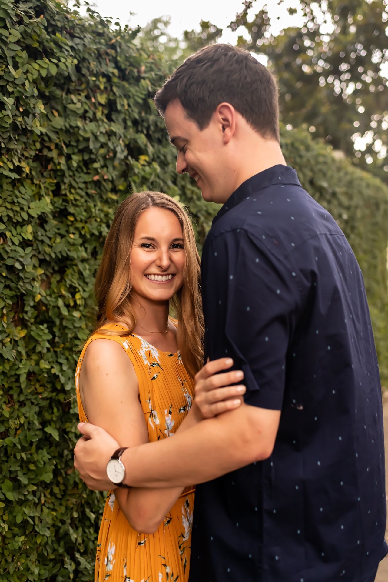 A couple embraces and smiles in front of a hedge during a warm outdoor photo session.