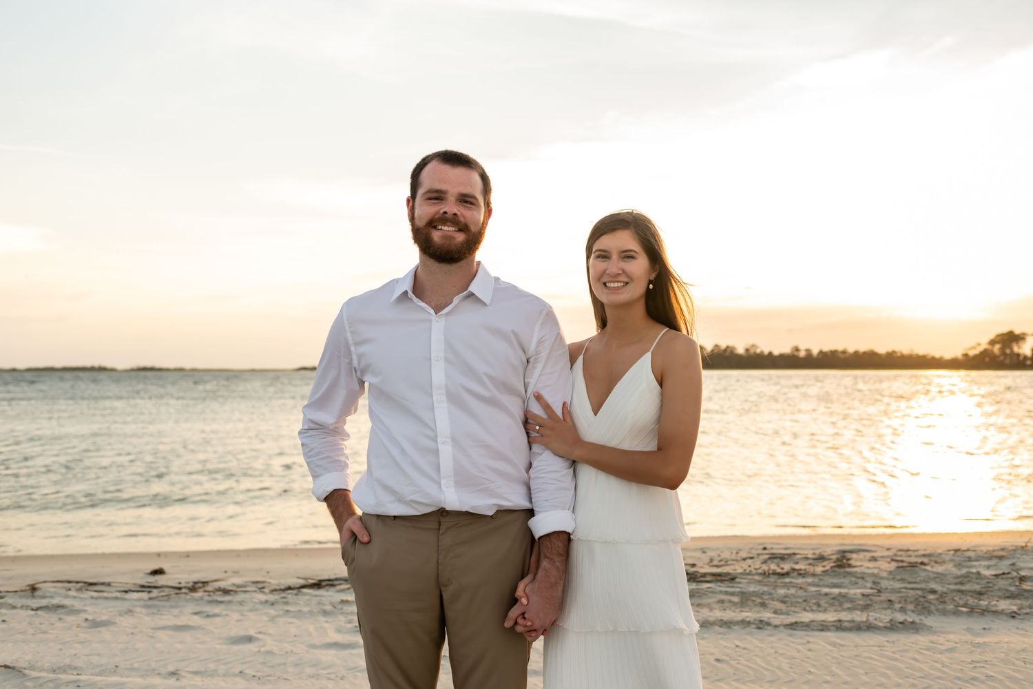 A couple poses together on a sunlit beach at sunset wearing white and tan casual attire.