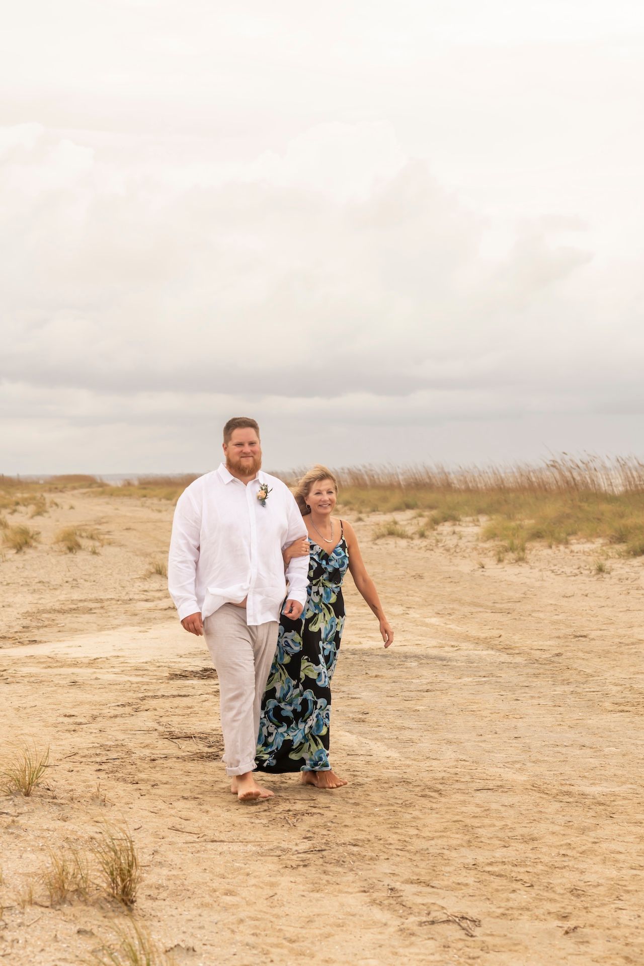 A couple takes a romantic stroll together along a sandy beach path with sea oats and cloudy skies in the background.