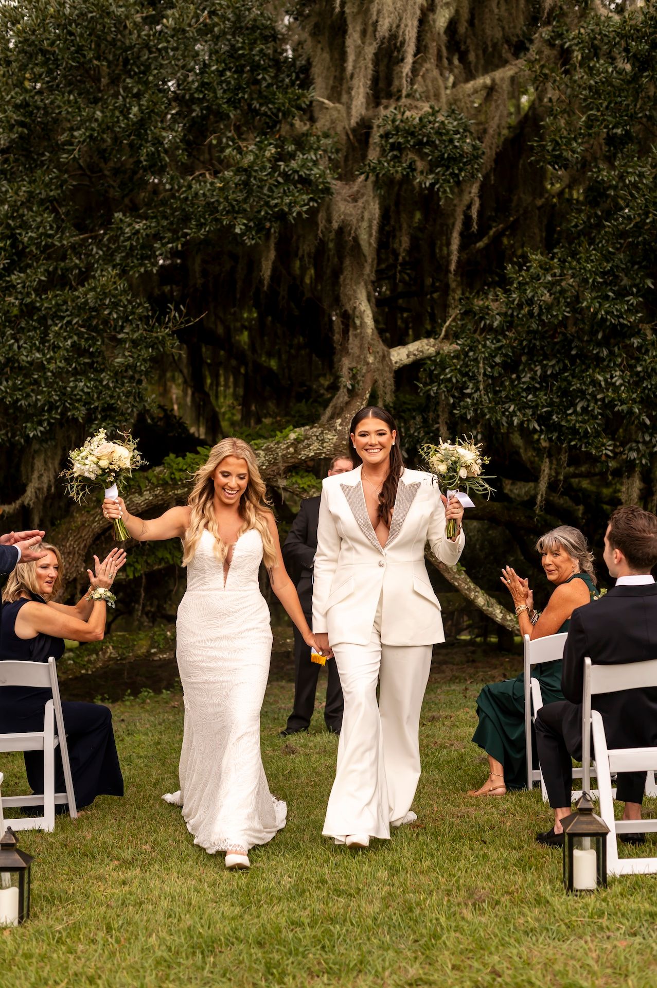People in white dresses celebrate outdoors at a wedding ceremony with white chairs and hanging moss trees in the background.