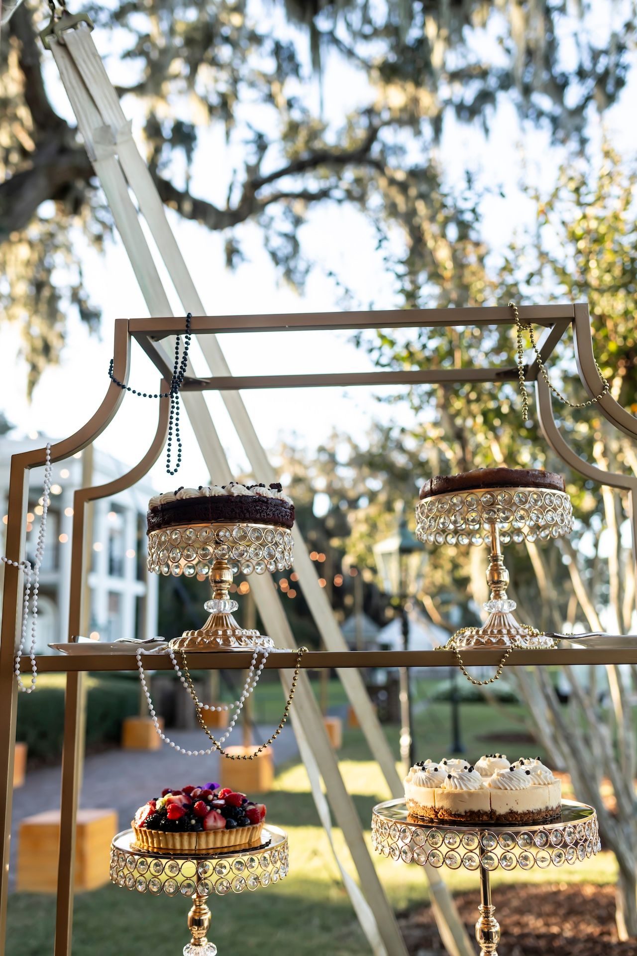 Crystal embellished cake stands with decorative chains displayed on an elegant tiered metal frame at an outdoor event setting.