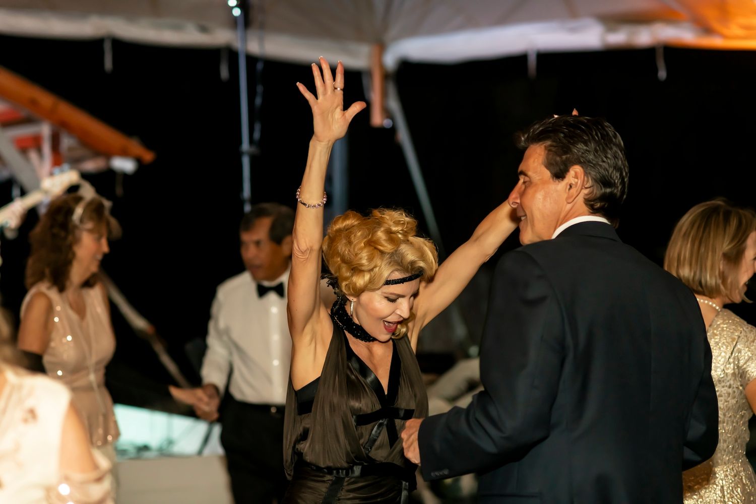 People dance and celebrate with raised hands on a dark dance floor at a party or wedding reception.