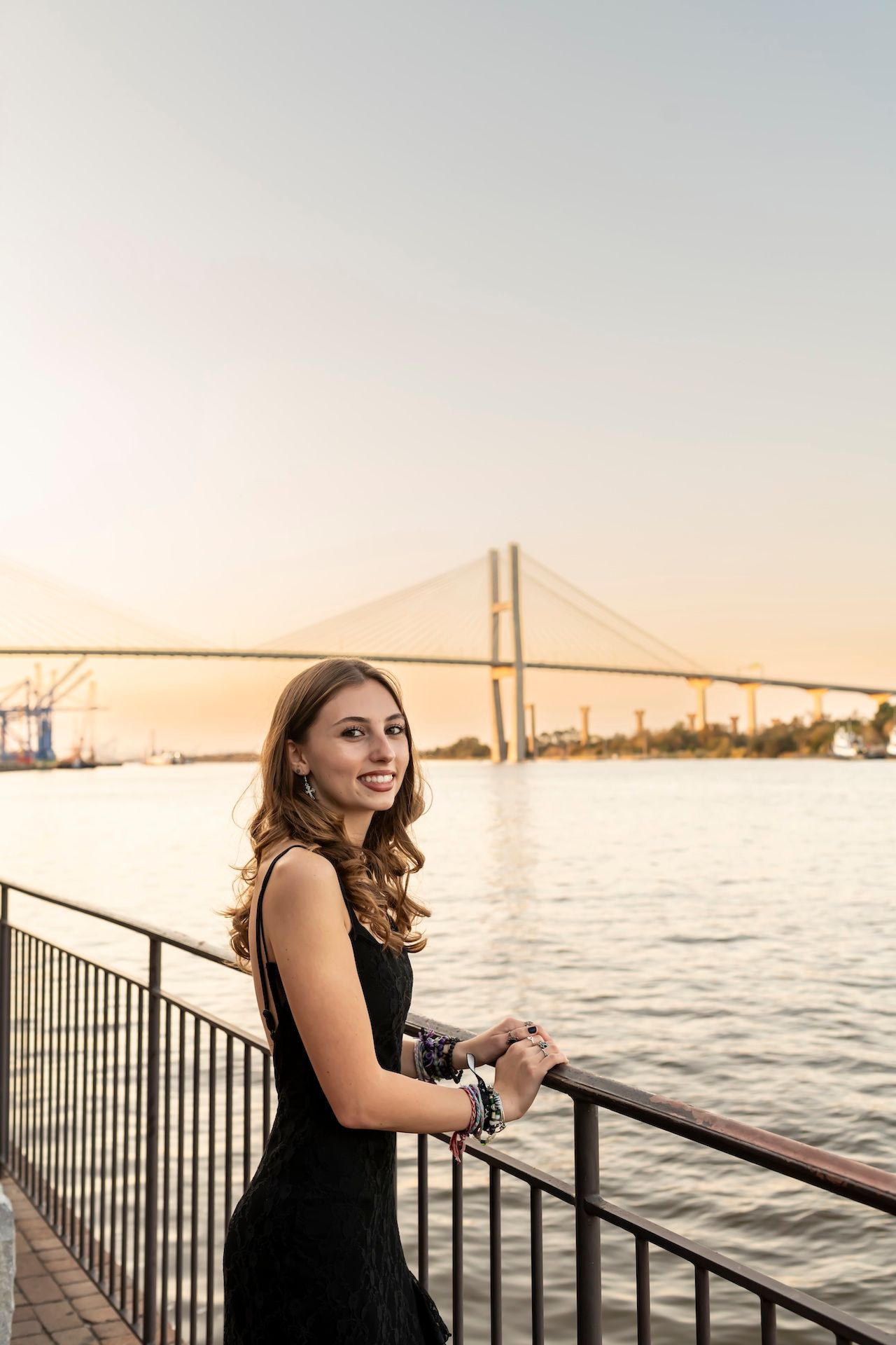 A person in a black dress stands at a railing overlooking a river with a suspension bridge at sunset.