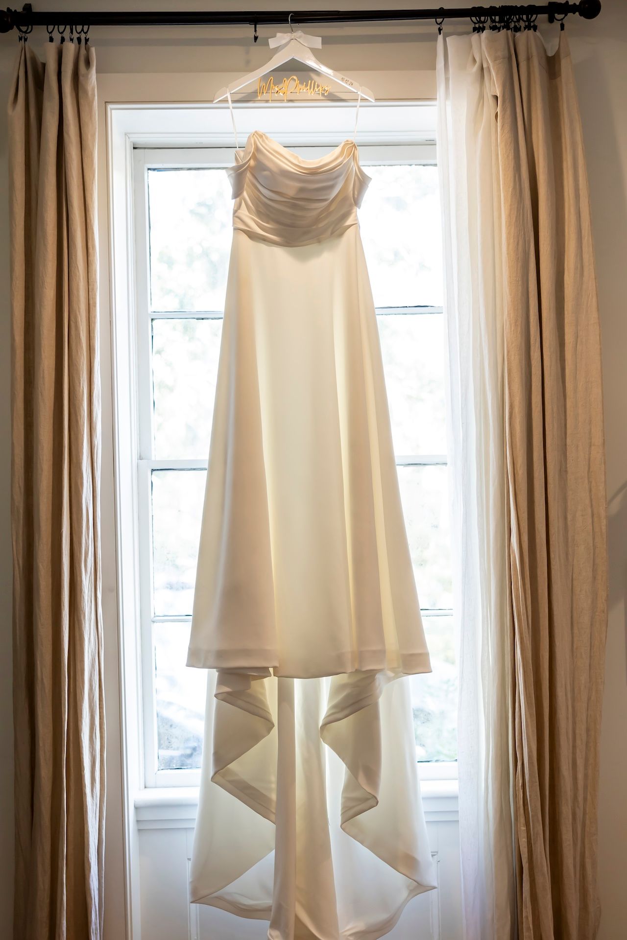 A white wedding gown hangs elegantly in front of a window with beige curtains.