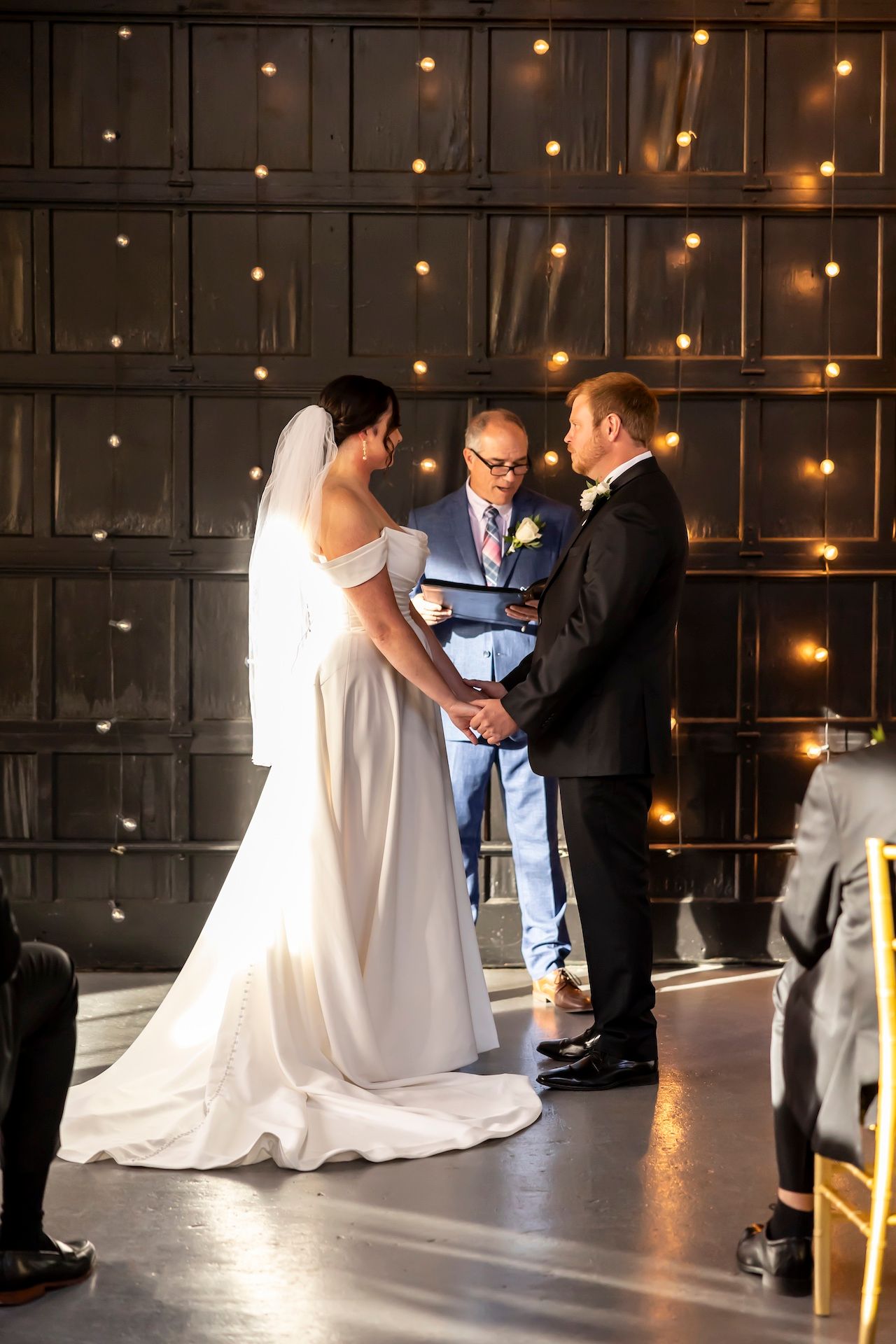 Couple stands before an officiant during their wedding ceremony against a dark wall illuminated with soft string lights.