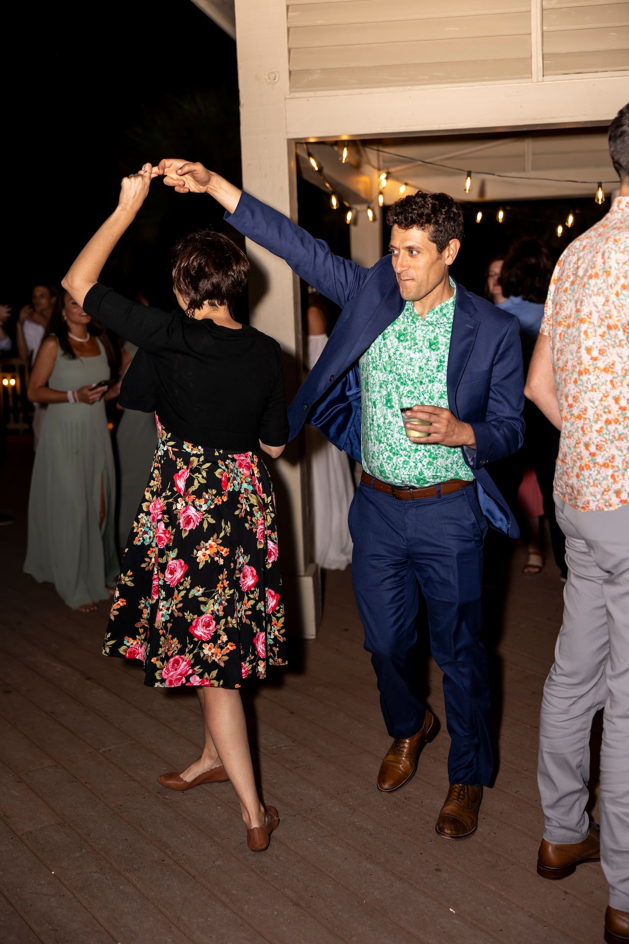 A couple dancing at a party, with the woman wearing a floral dress and the man in a blue suit and patterned shirt.