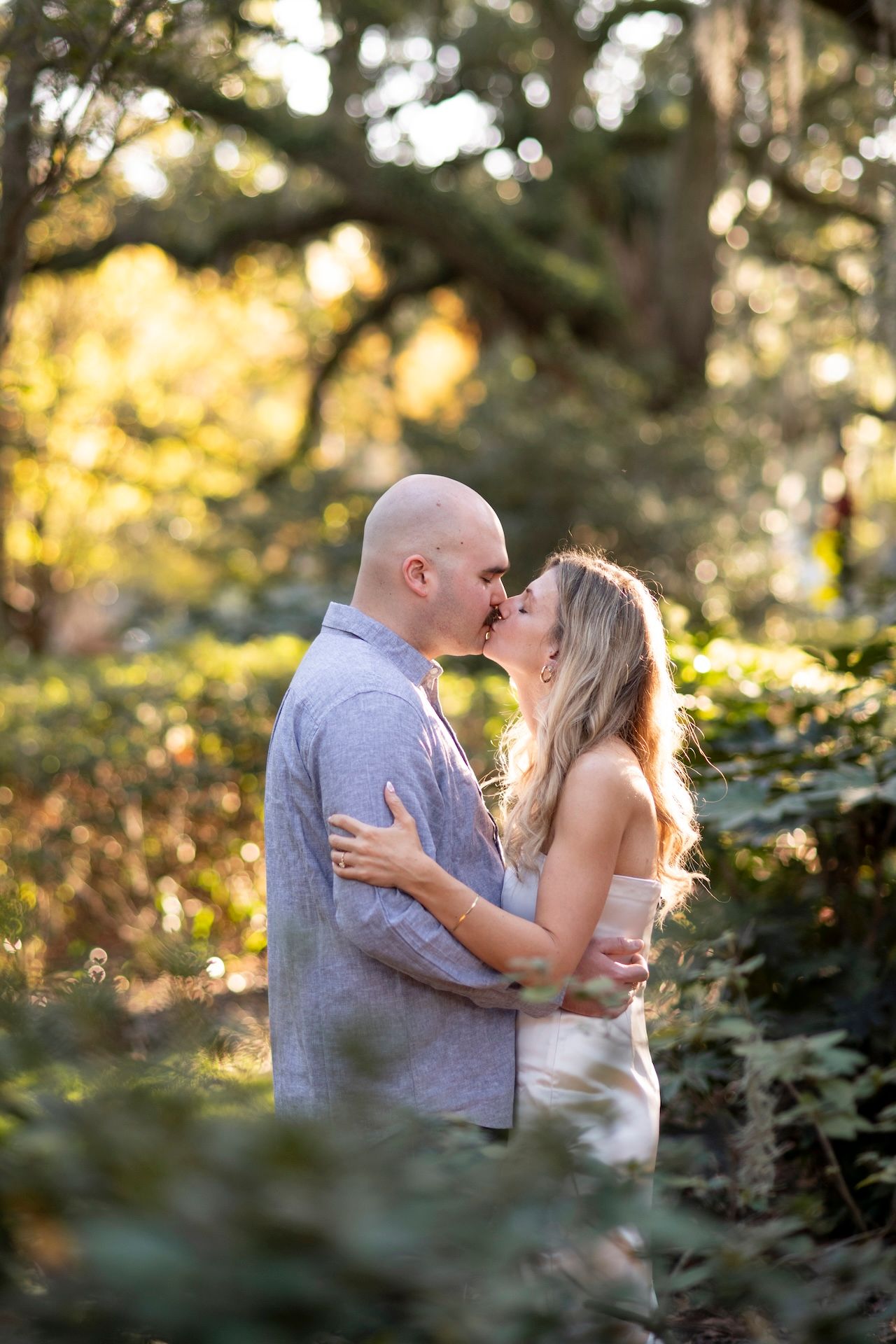 A romantic sunset engagement photo captures a couple sharing a kiss in a dreamy forest setting with golden light filtering through.