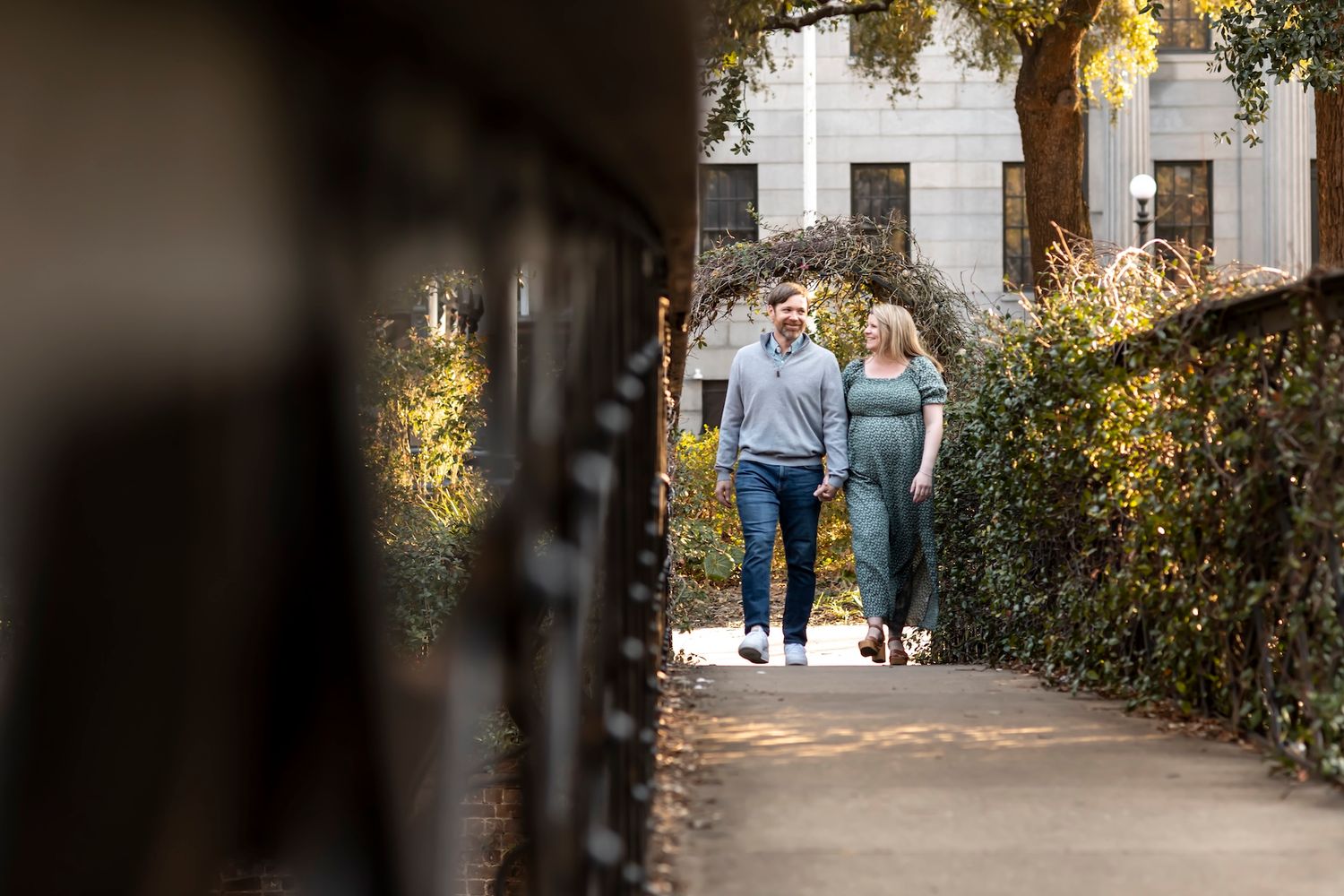 A couple strolls down a sunlit garden path between stone buildings with lush greenery and bushes lining the walkway.