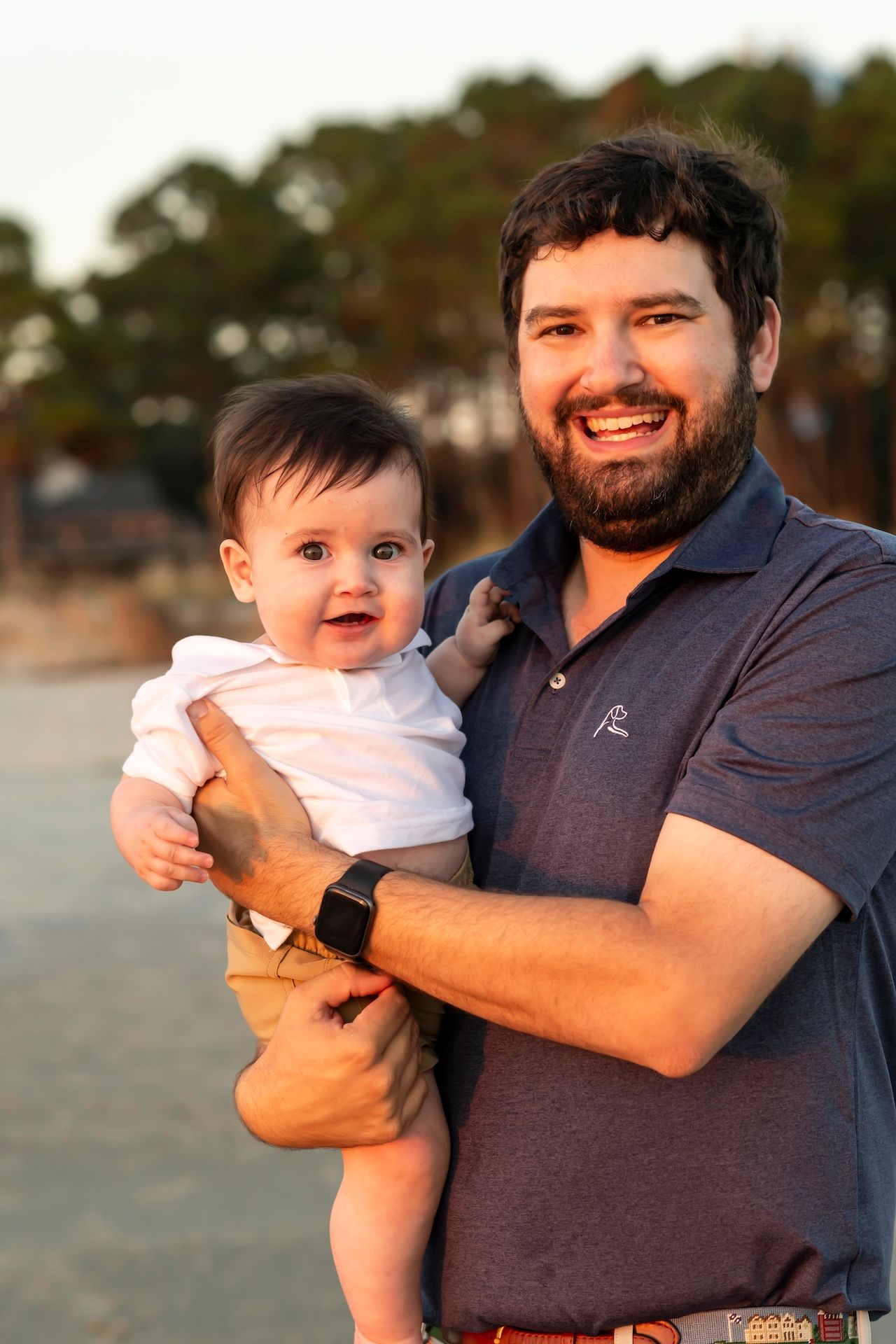A bearded parent in a navy polo shirt holds a happy baby in white against a sunset beach backdrop.