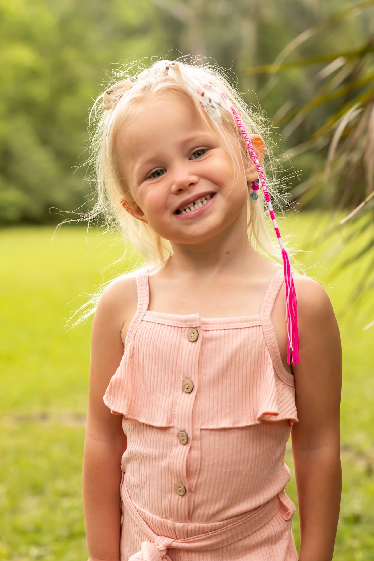 Child wearing a pink button-up sundress smiles brightly in a garden setting with green foliage in the background.
