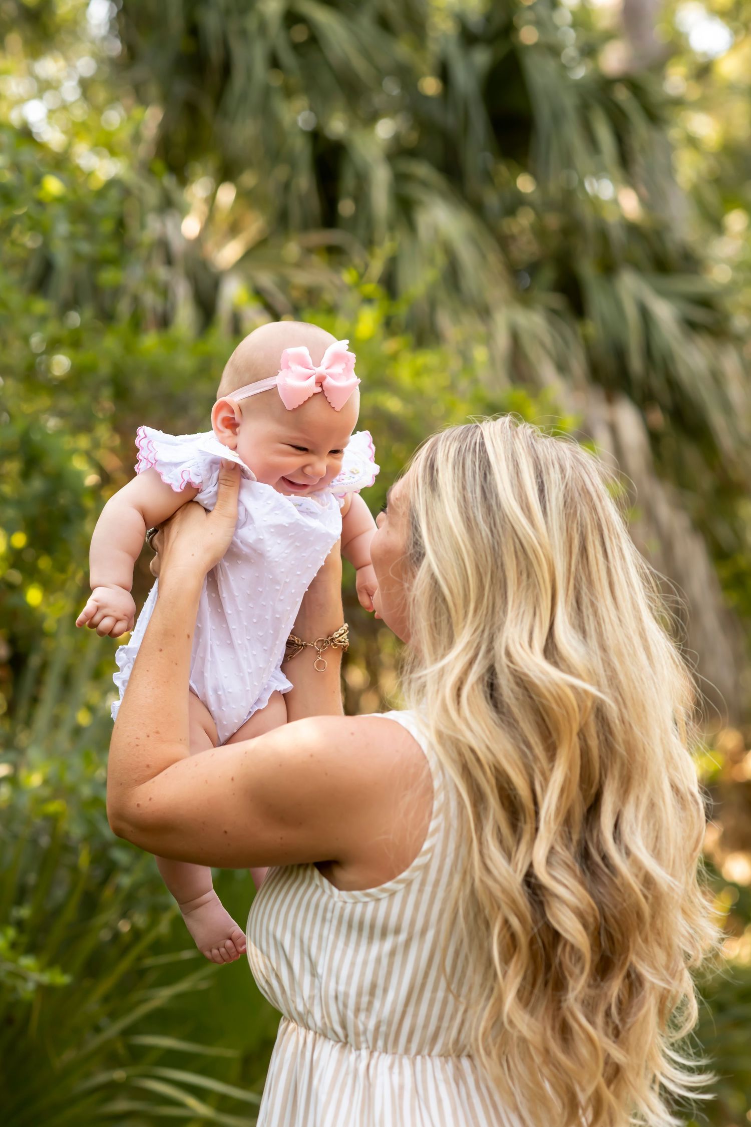 A woman in a striped dress lifts a baby wearing pink and white in a lush tropical garden setting.