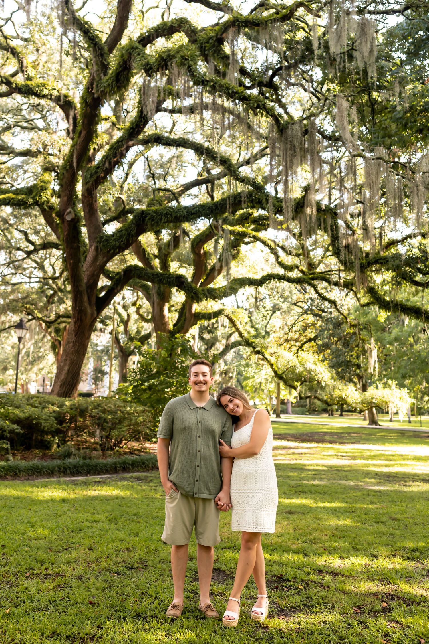 A couple stands together beneath moss-draped oak trees in a sunlit southern park setting.