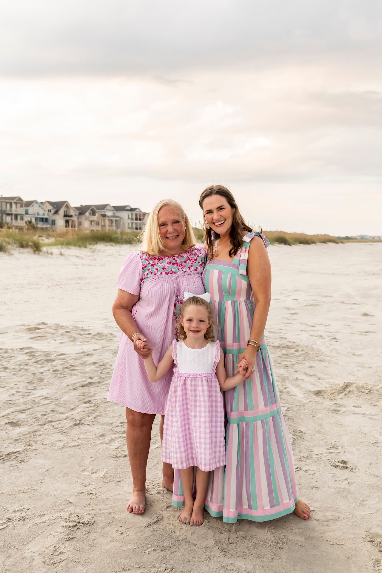 Three people wearing coordinating pink, white and blue dresses pose together on a sandy beach at sunset.