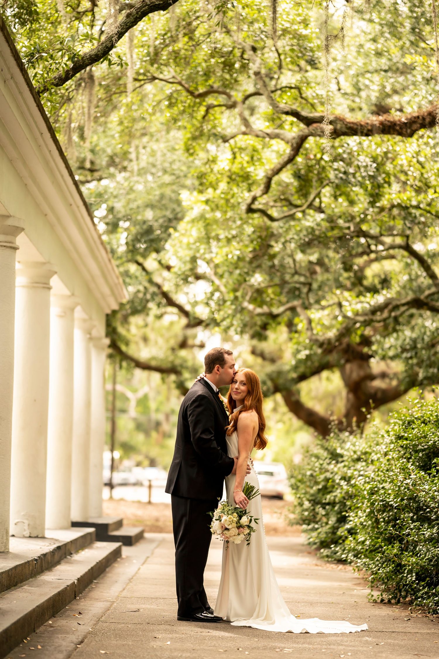 A couple shares an intimate moment under a tree-lined colonnade with white columns and lush greenery in the background.