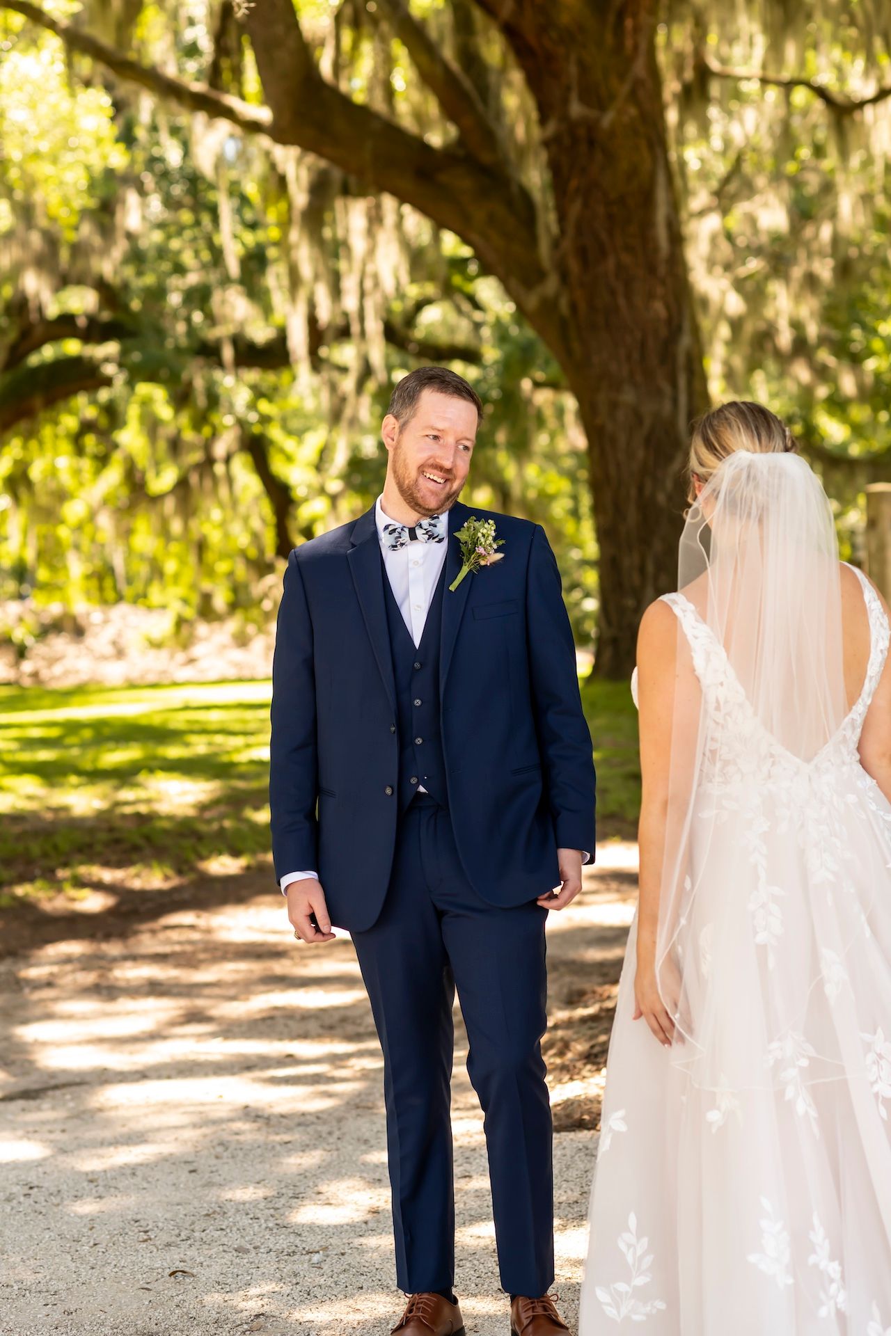 A first-look moment between a couple in formal wedding attire under Spanish moss-draped trees in a sunlit garden setting.