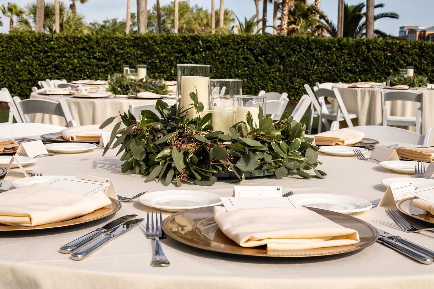 Elegant outdoor table setting with white linens, gold charger plates and lush greenery centerpieces at garden reception.