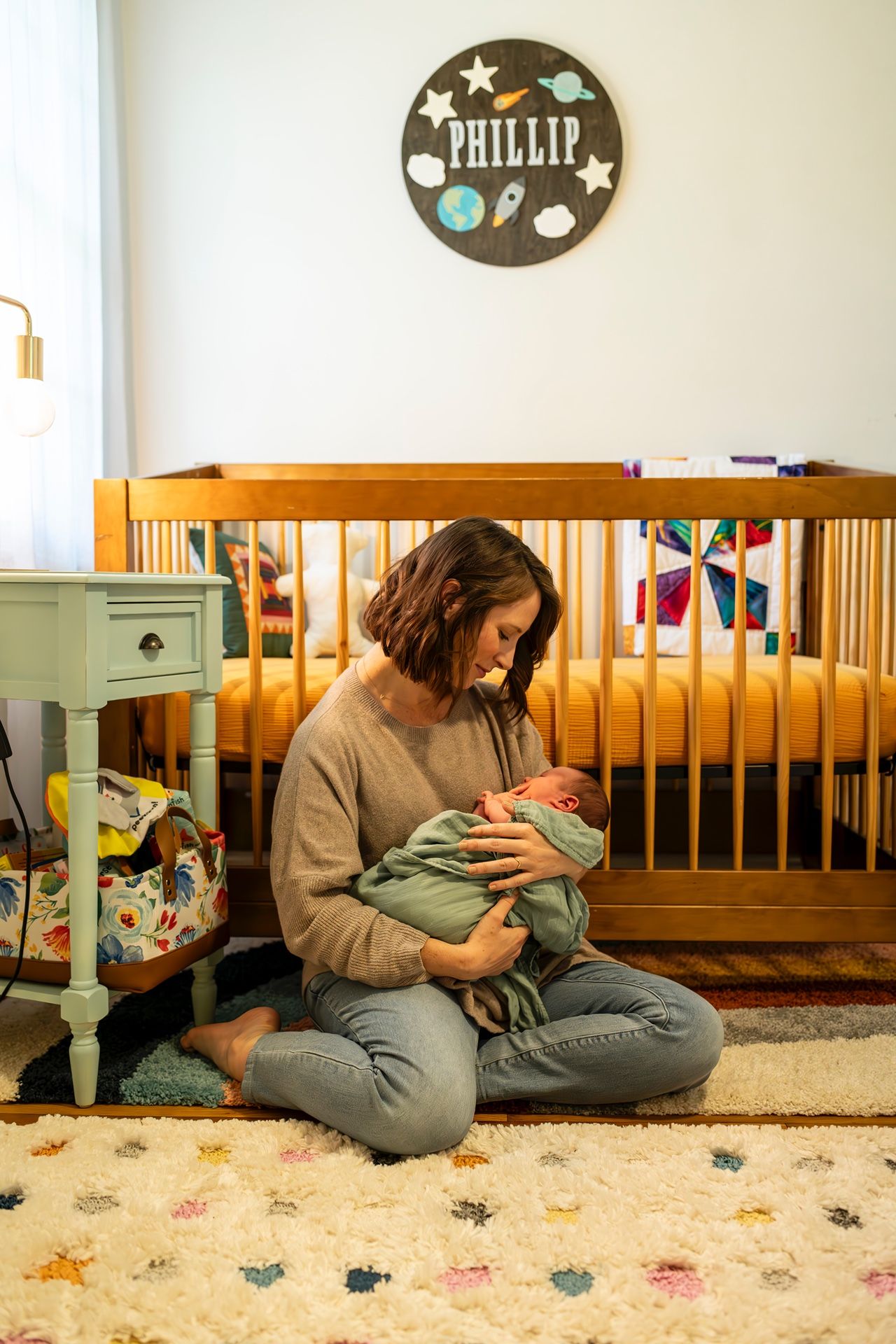 Person in beige sweater sitting on nursery floor with infant beside wooden crib and decorative wall sign reading Phillip.