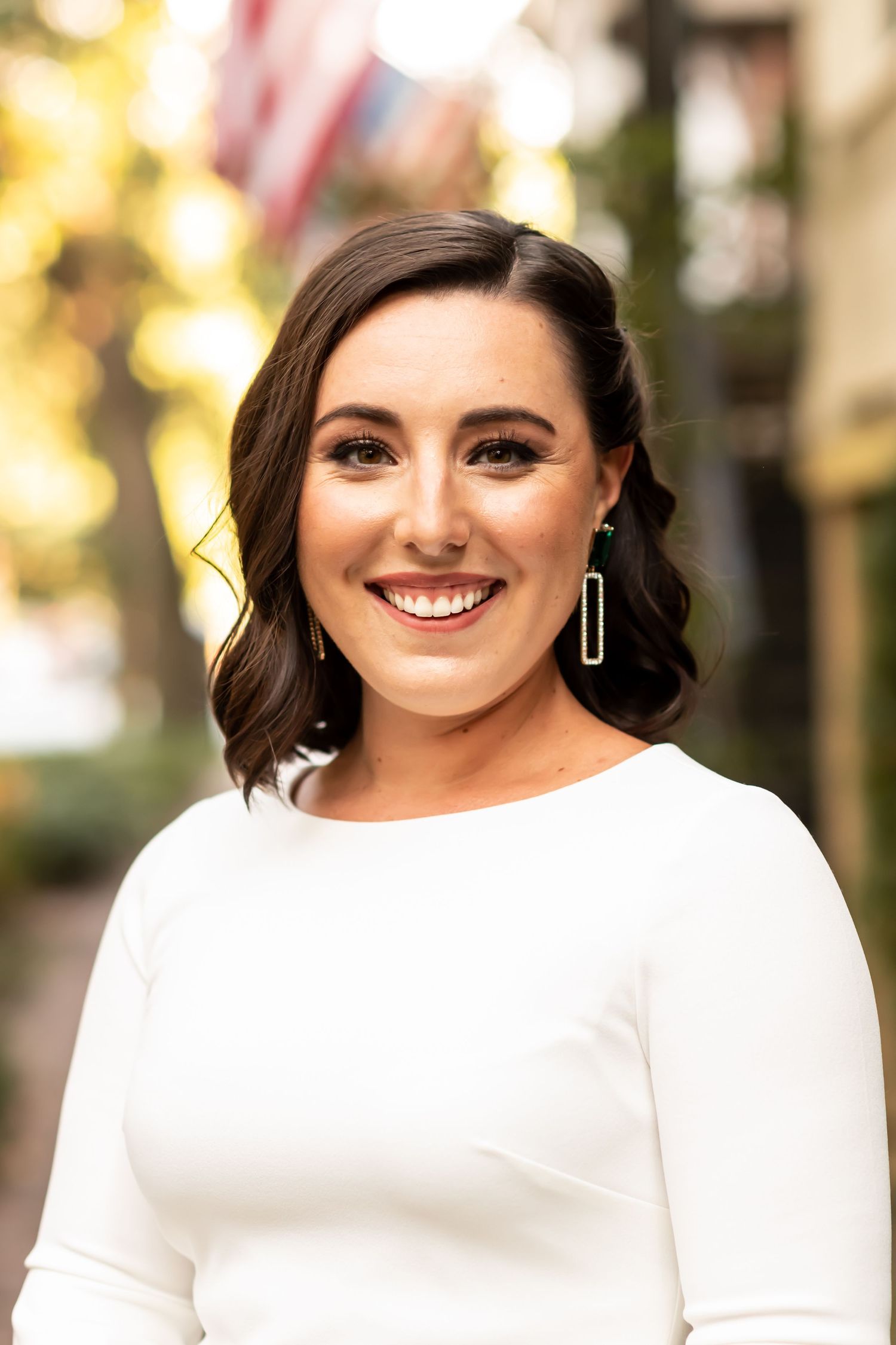 Professional headshot portrait of someone in a white long sleeve dress against a blurred outdoor background.