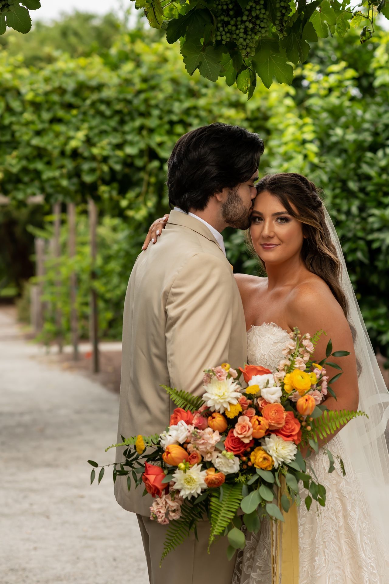 A couple embraces in a garden path while holding a vibrant bouquet of coral, yellow and white flowers arranged with ferns.