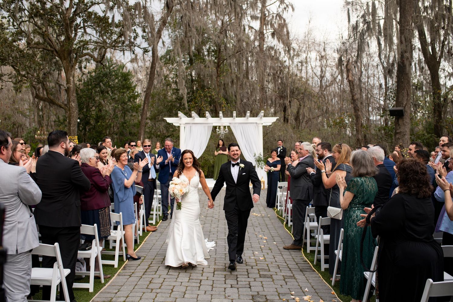 Bride and groom walk down outdoor aisle after wedding ceremony, guests cheering, white pergola backdrop.