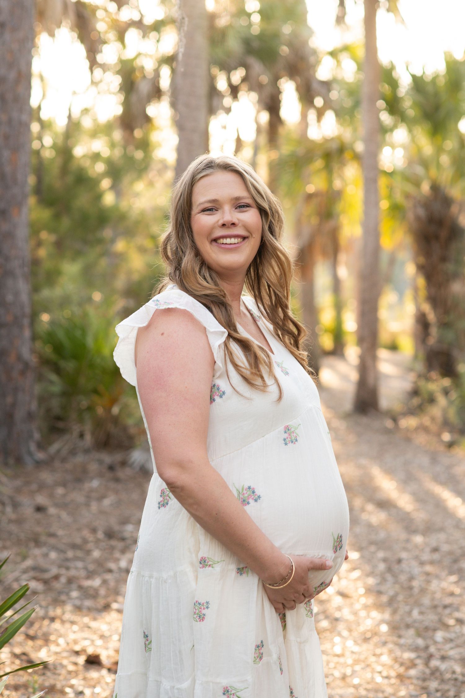 Smiling pregnant woman in white floral dress holding baby bump among sunlit trees.