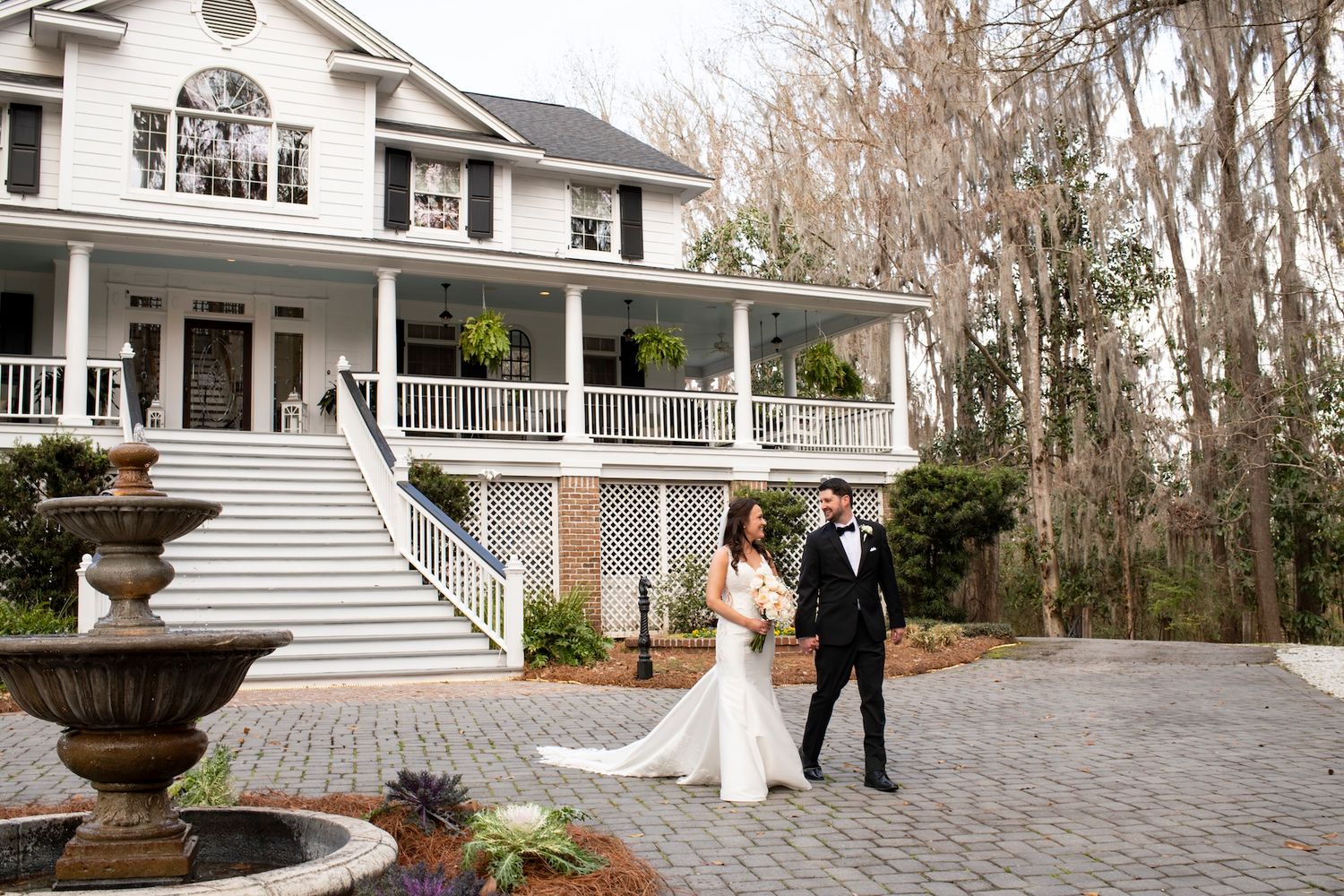 Bride and groom walk hand in hand on a cobblestone driveway in front of an elegant white Southern mansion.
