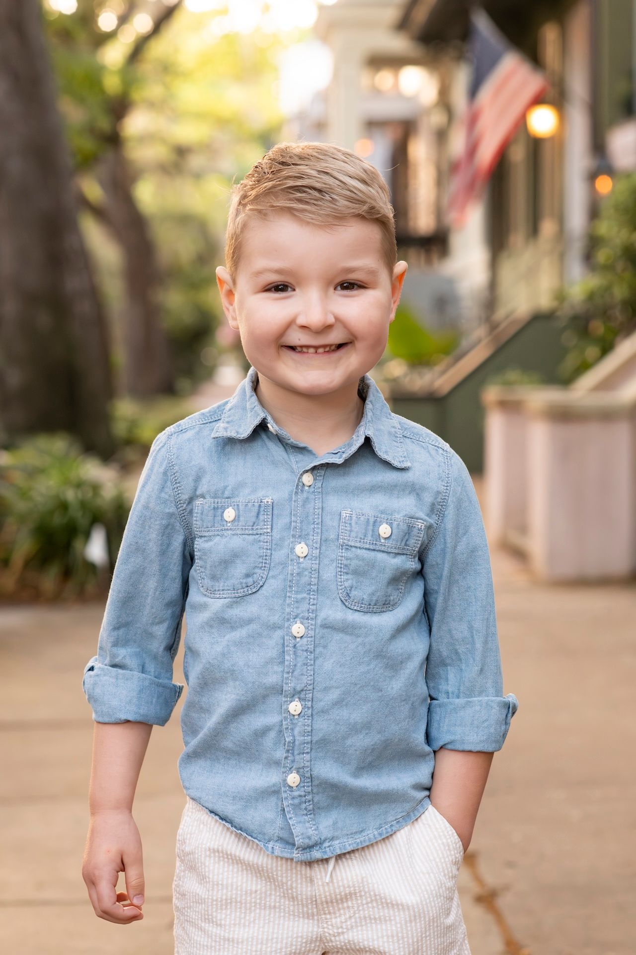 Smiling young boy in a blue chambray shirt and white pants standing outdoors near an American flag.