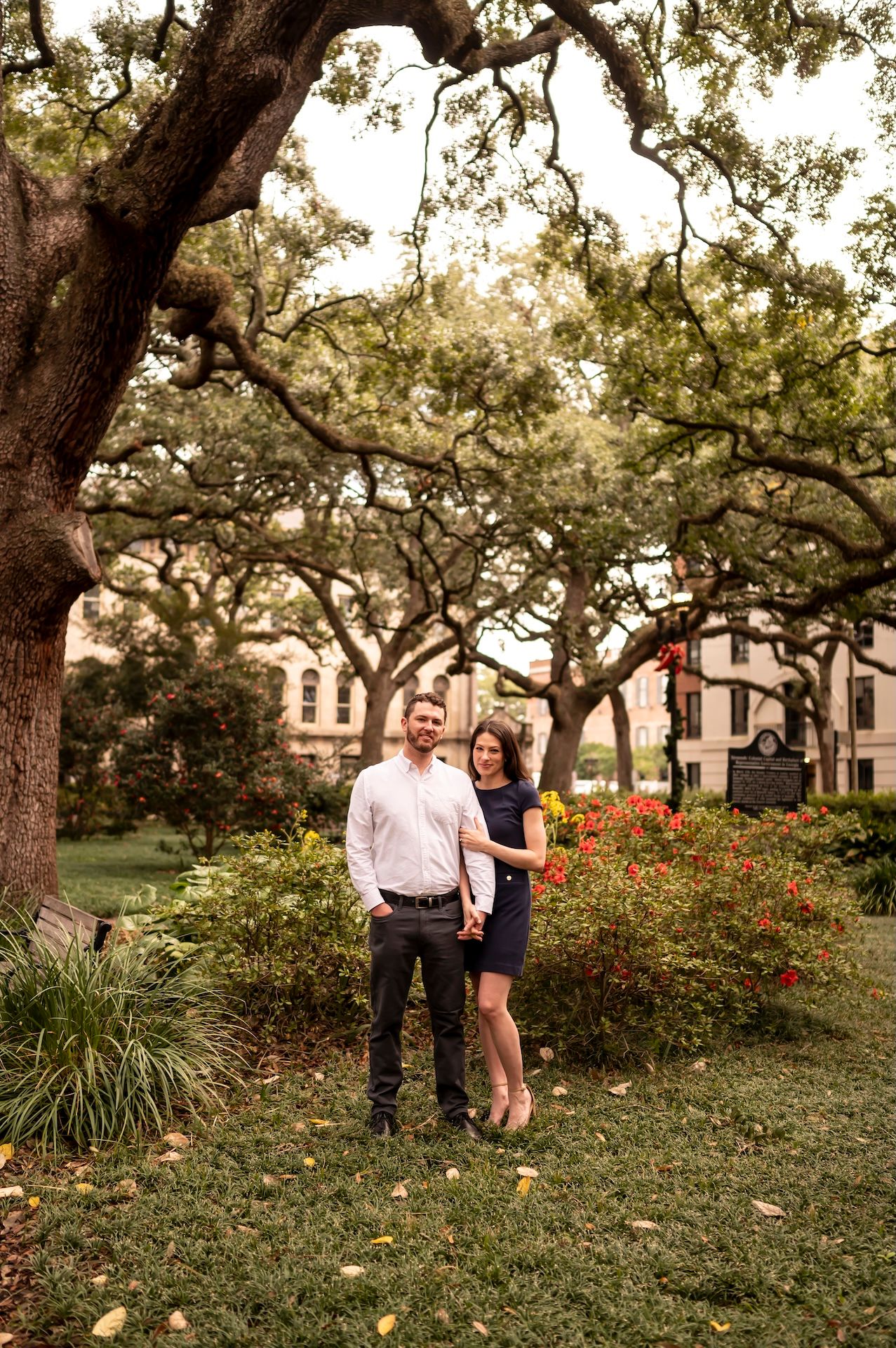 A couple stands together under large oak trees in a garden setting with blooming azalea bushes at sunset.